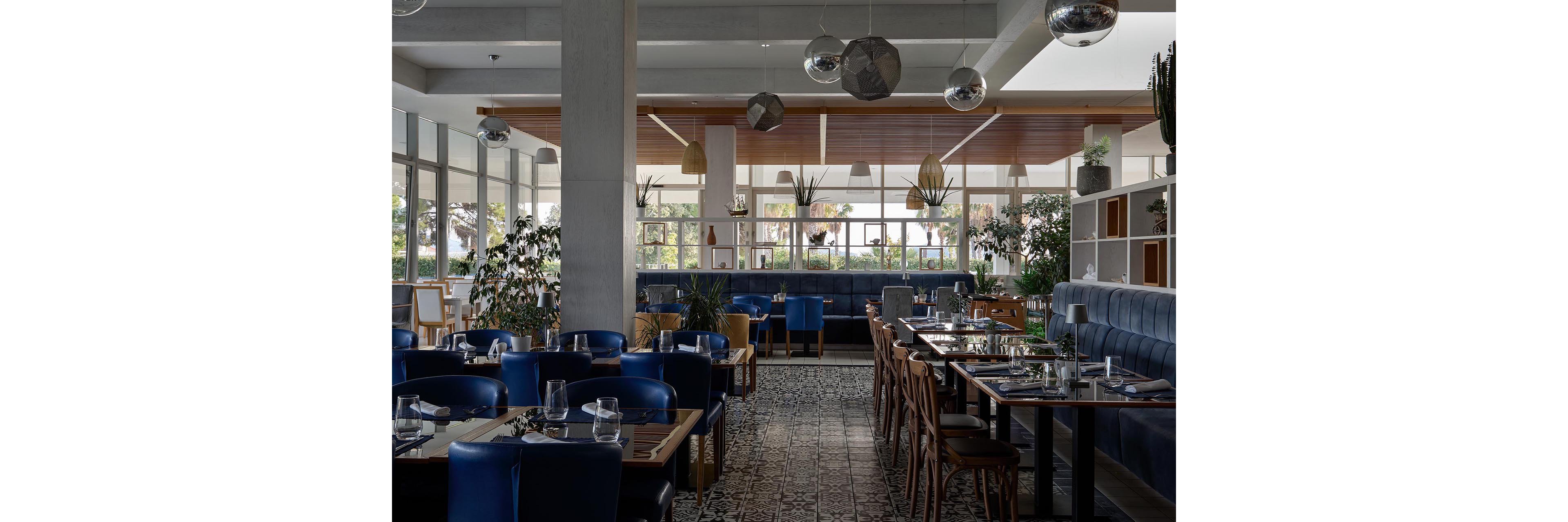 Contemporary restaurant dining room with neatly set tables and blue upholstered seating. Large windows let in natural light and offer views of greenery outside. Decorative plants, patterned floor tiles, and hanging light fixtures create a stylish, relaxed atmosphere. The space appears ready for guests, with glassware and table settings arranged in an orderly fashion.