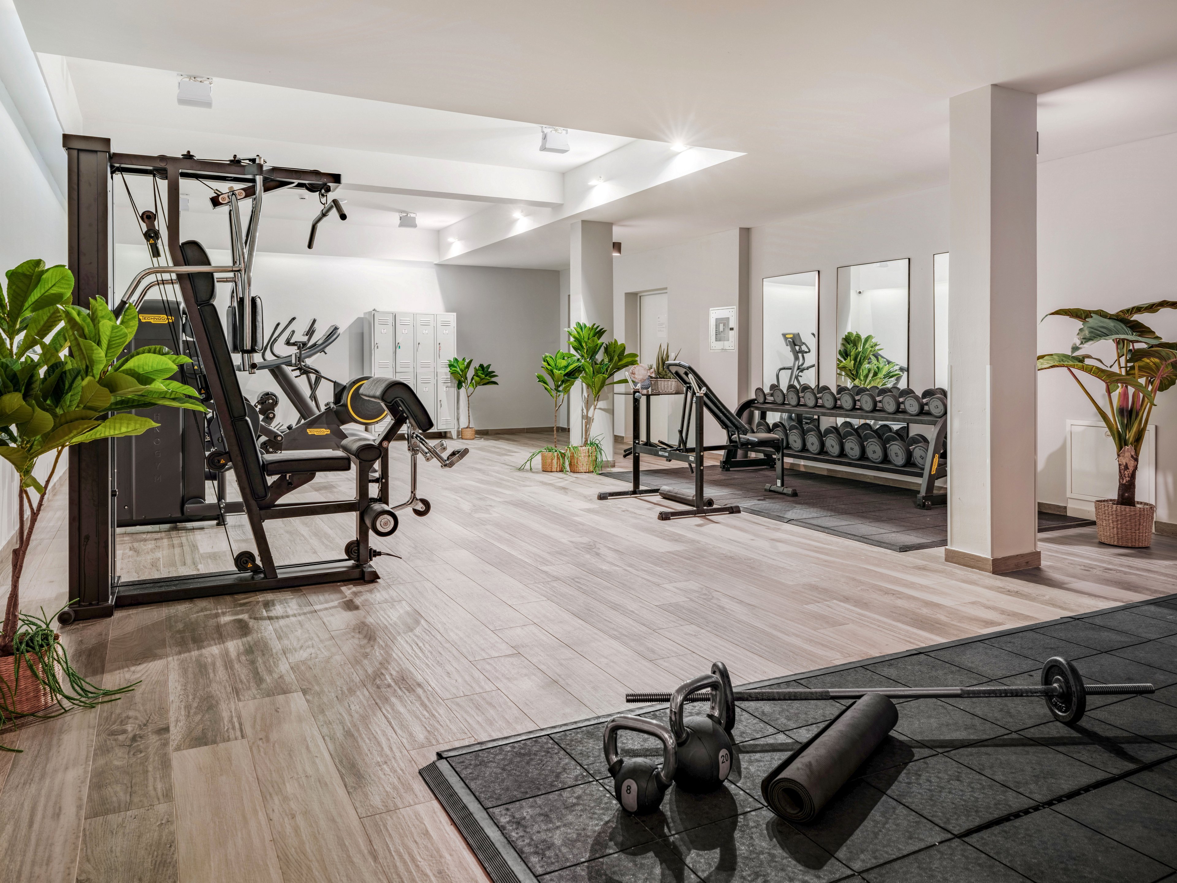 A spacious modern gym features a variety of strength training machines and free weights neatly arranged in a bright, open room. Dumbbells are lined up on a rack beside benches, while a multi-gym station stands near the wall. A yoga mat, kettlebells, and a foam roller are placed on the floor in the foreground, suggesting functional training or stretching. Large mirrors and potted plants add to the clean, contemporary atmosphere of the fitness space.