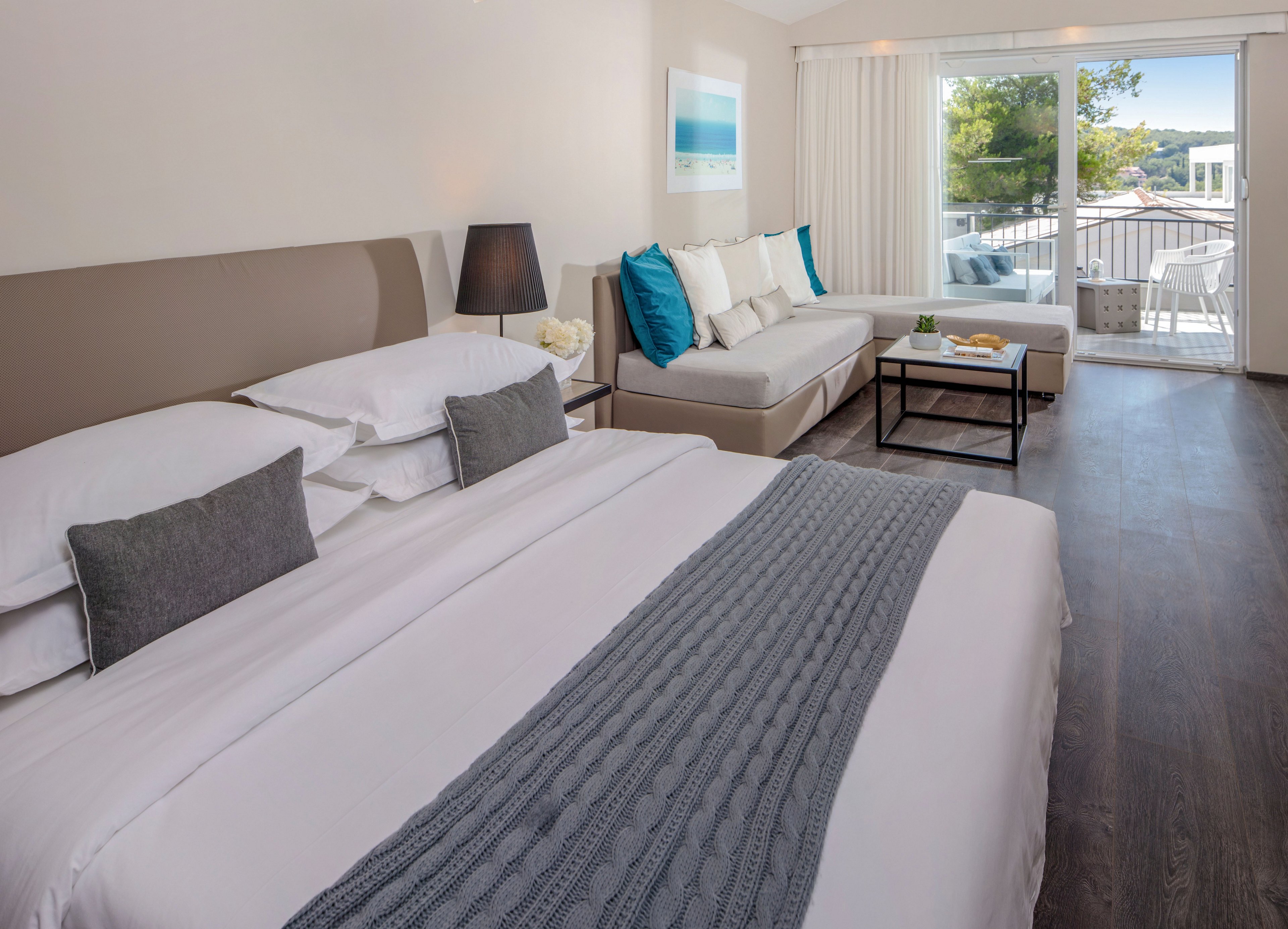 A contemporary hotel bedroom features a large neatly made bed with white linens and a gray knitted throw. Beside the bed, a sofa with white cushions and turquoise accent pillows faces a low coffee table. A floor lamp and framed seascape artwork add decorative touches to the neutral-toned interior. Large sliding glass doors open to a sunny balcony with outdoor seating and a view of trees and nearby buildings.
