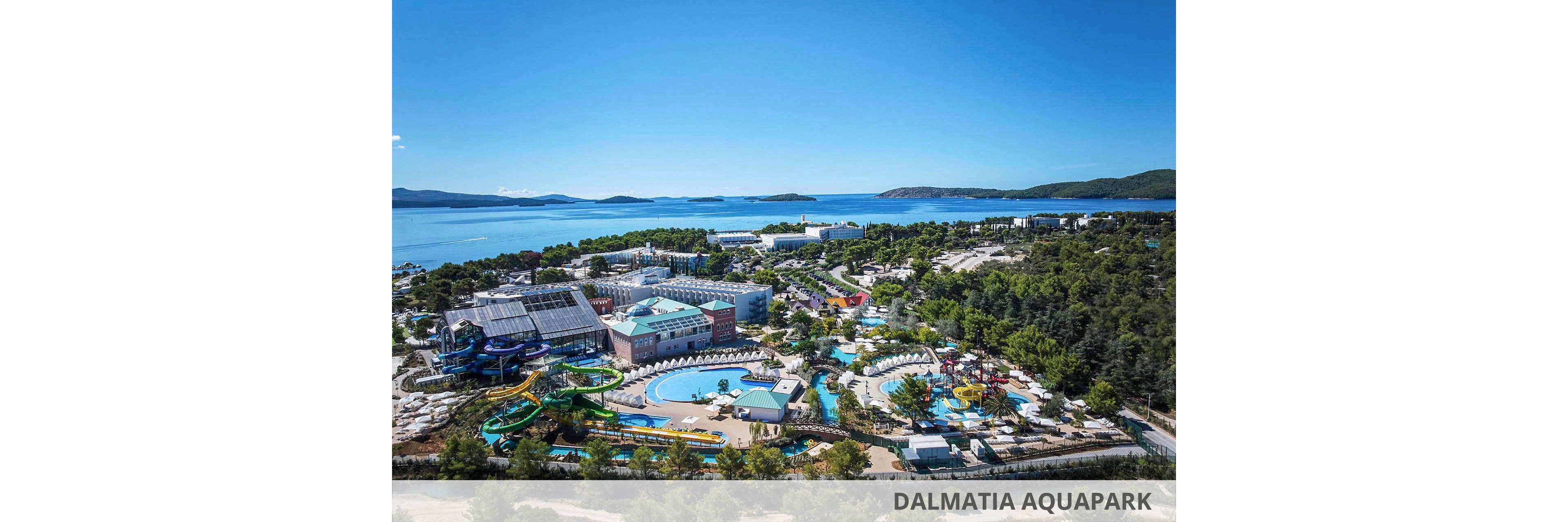 An expansive coastal resort with a large water park is captured from an elevated viewpoint. Multiple pools, winding water slides, and modern buildings are surrounded by dense green trees. The resort sits along a calm blue bay with islands and distant hills on the horizon. Clear skies and bright sunlight create a vibrant, summery atmosphere ideal for vacation and leisure.