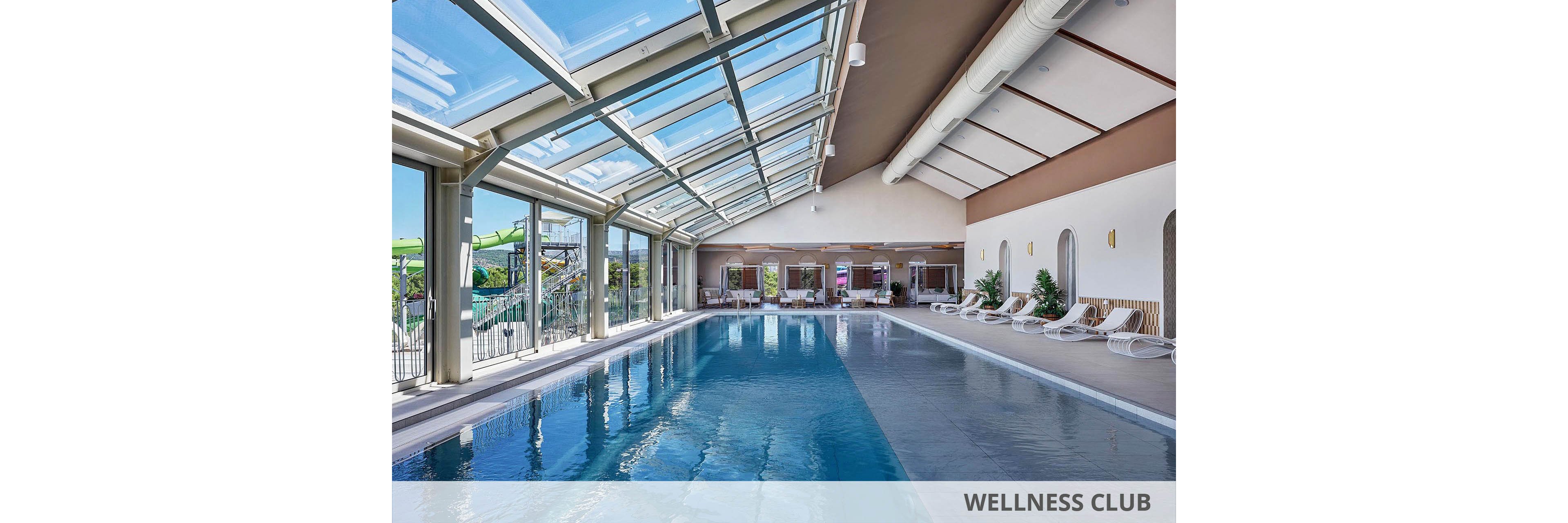A spacious indoor swimming pool stretches along a sunlit room with a large glass skylight roof. Floor-to-ceiling windows line one side, offering views of an outdoor water play area. White lounge chairs and potted plants are arranged neatly along the opposite wall, creating a relaxing resort atmosphere. The clear blue water and clean architectural lines emphasize a modern, tranquil leisure space.