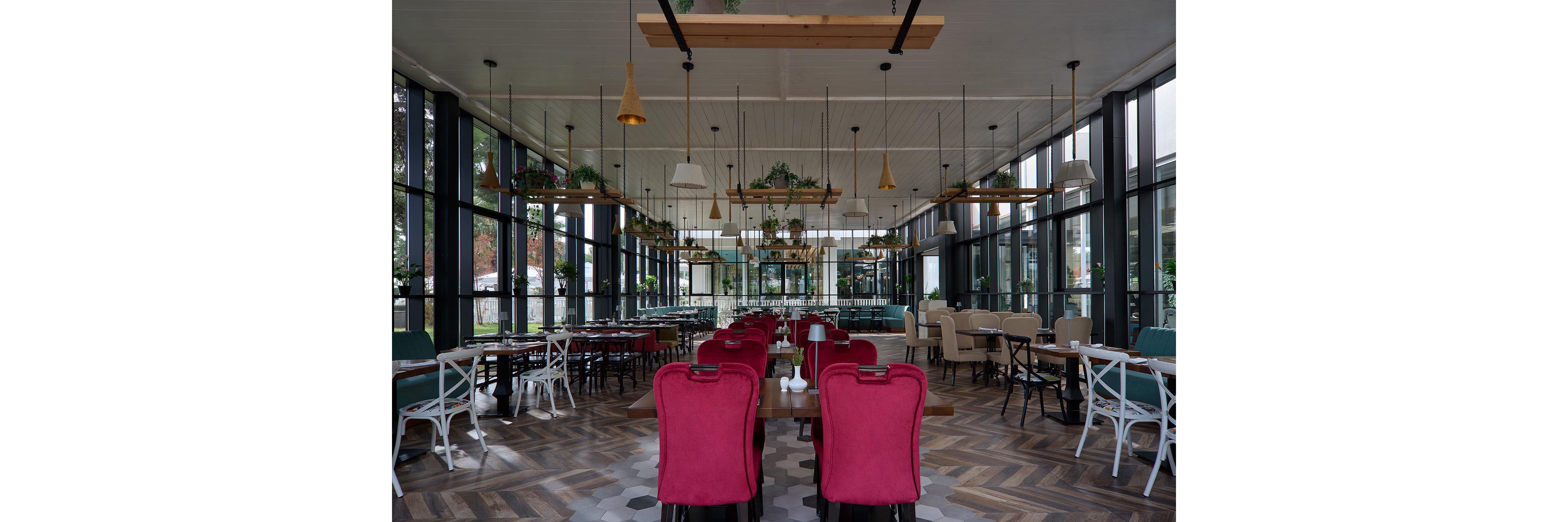 A contemporary restaurant interior features rows of red upholstered chairs and wooden tables arranged neatly in the center. Large floor-to-ceiling windows surround the space, allowing natural light to illuminate the dining area. Hanging pendant lights, potted plants, and geometric patterned flooring add stylish decorative elements. The restaurant appears empty, emphasizing the spacious layout and modern design details.