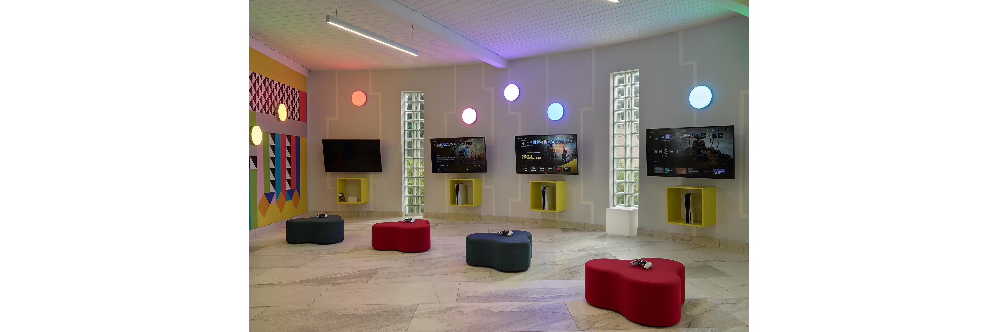 A modern gaming lounge features multiple flat-screen televisions mounted on a light-colored wall. Each screen is paired with a game console and controller, with curved ottomans placed in front for seating. Circular wall lights in different colors and geometric wall art add a playful, vibrant atmosphere. The space has a clean, minimalist design with bright accents and ample open floor area.