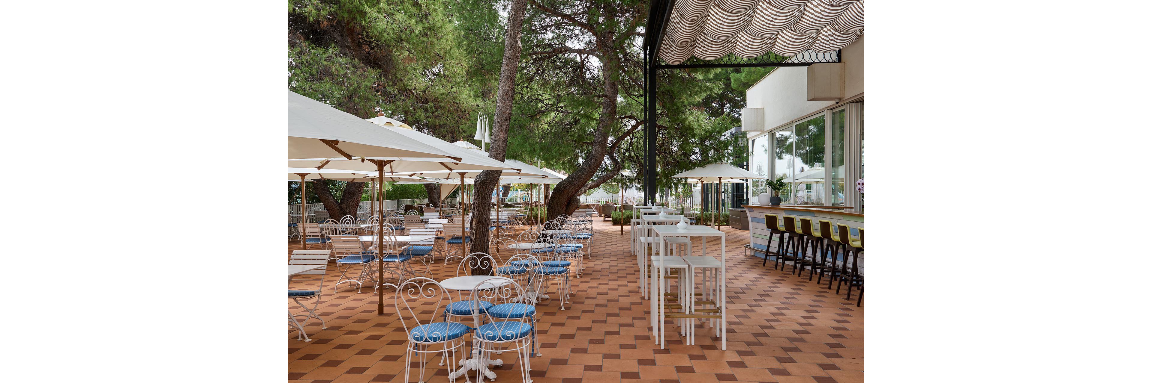 An outdoor café terrace features white metal chairs with blue cushions arranged around small round tables. Large white umbrellas provide shade along the tiled patio, which is lined with tall trees creating a natural canopy. On the right side, a bar counter with high stools faces large glass windows of the adjacent building. The scene is calm and inviting, suggesting a relaxed dining or coffee setting in a warm climate.