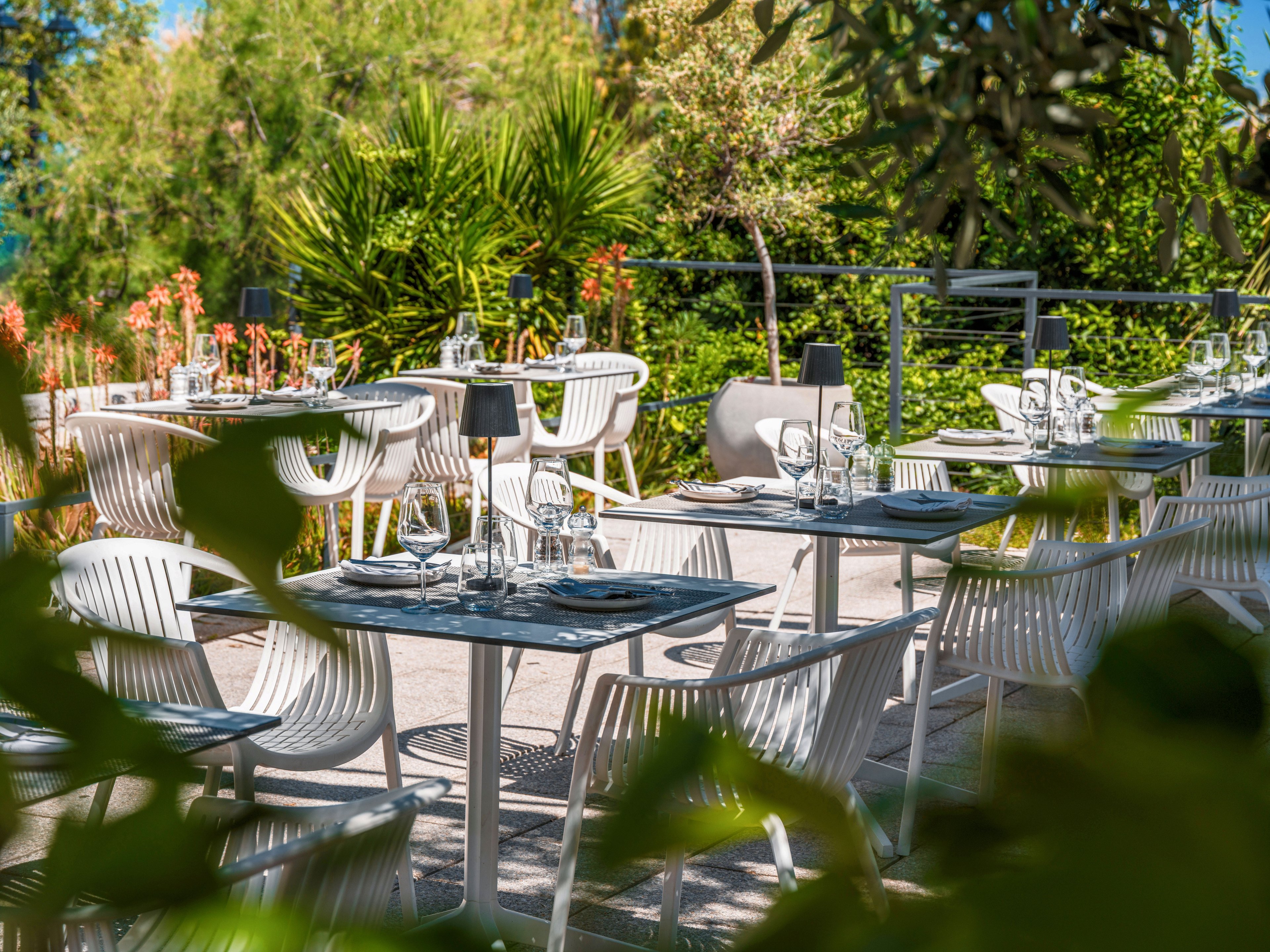 An inviting outdoor restaurant terrace is set with modern white chairs and neatly arranged tables. Each table features glassware, plates, and small black table lamps, ready for guests. Lush green plants and trees surround the seating area, creating a vibrant, natural backdrop. Bright sunlight enhances the relaxed, summery atmosphere of the dining space.