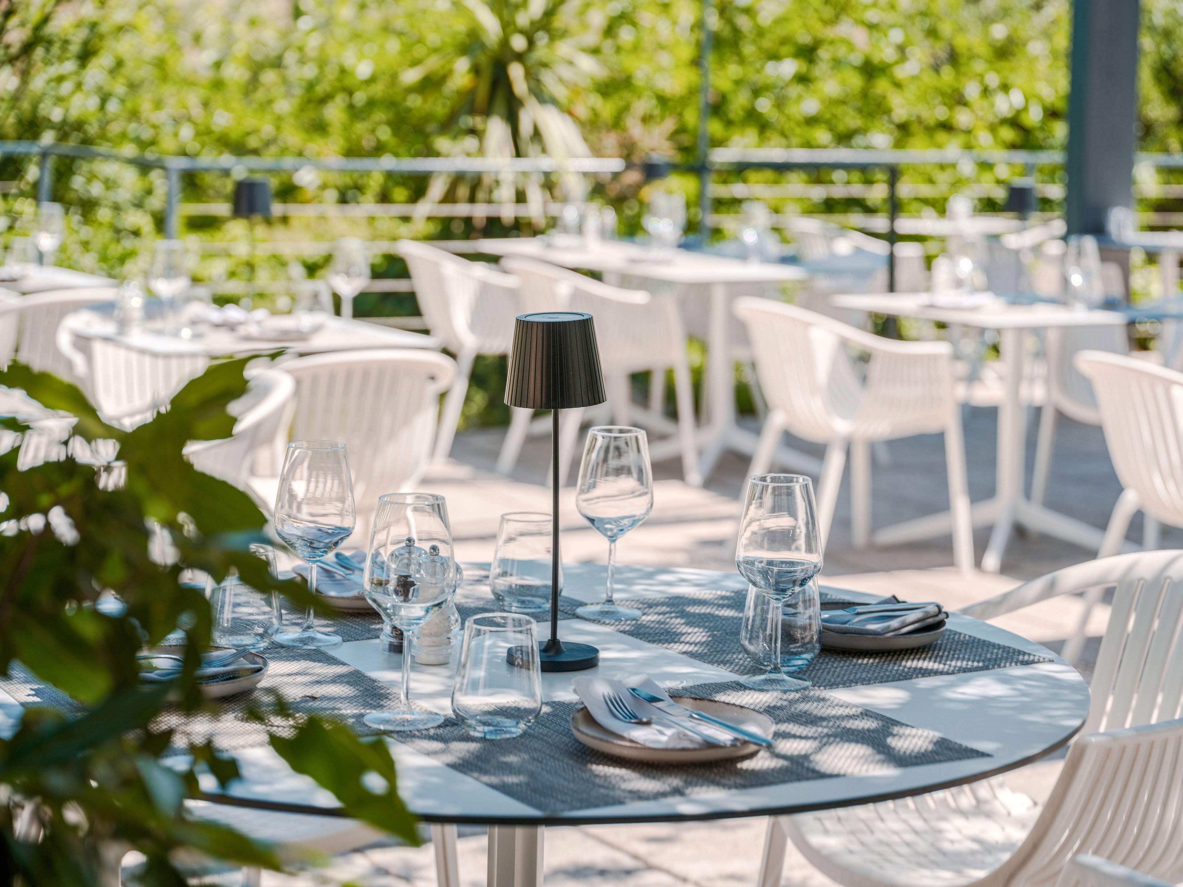 A round metal table is neatly set with wine glasses, water glasses, plates, and cutlery in an outdoor restaurant. White chairs surround the tables, arranged on a sunlit terrace with railings. Lush green plants and trees create a fresh, natural backdrop. The scene suggests a relaxed, elegant dining experience in warm daylight.
