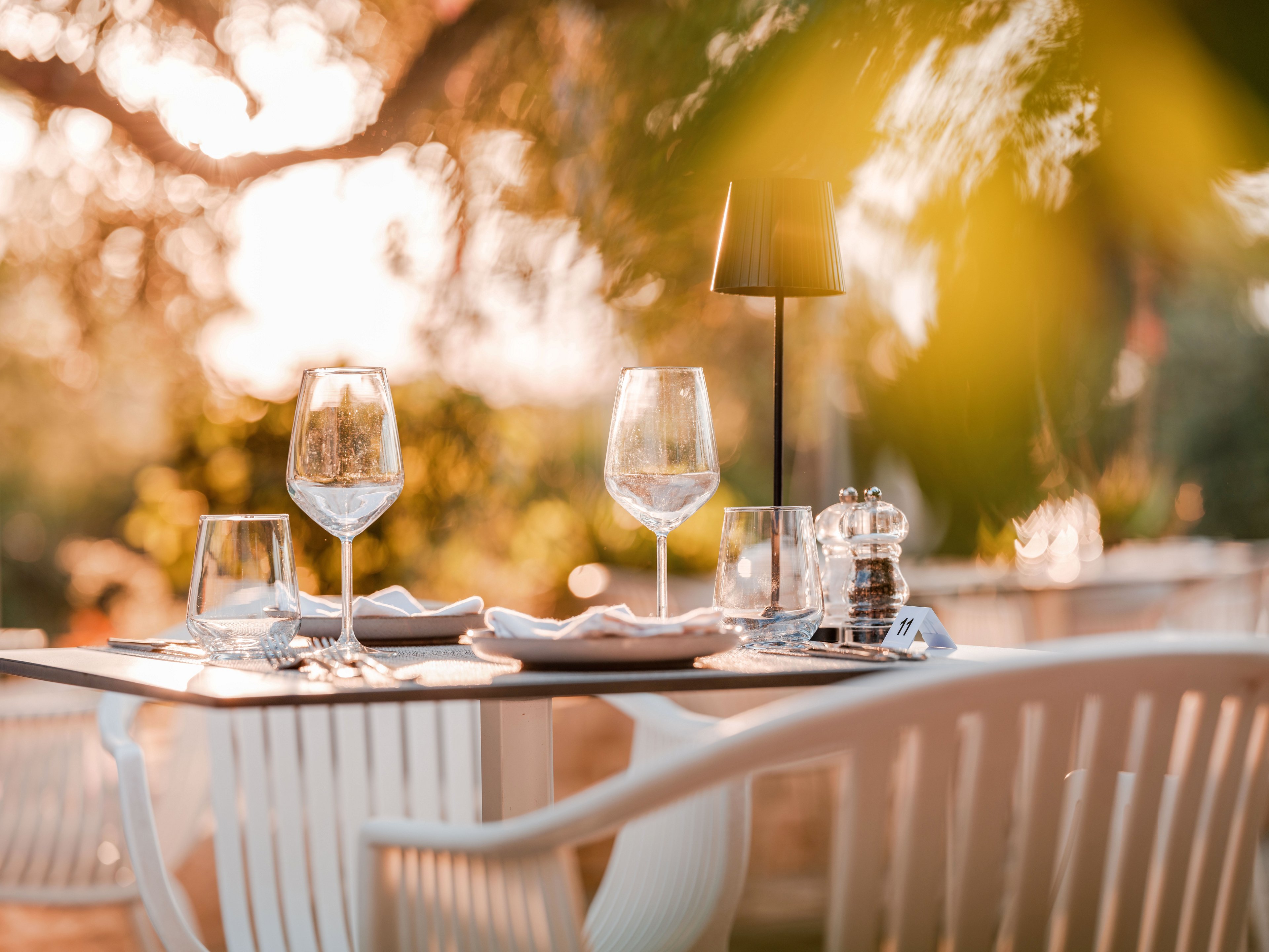 A neatly set outdoor dining table features empty wine glasses, water glasses, and folded napkins arranged on a smooth tabletop. A small modern table lamp and a pair of clear salt and pepper shakers add detail to the scene. White chairs surround the table, while soft golden sunlight filters through foliage in the background. The warm, blurred garden setting creates a romantic and relaxed atmosphere ideal for al fresco dining.