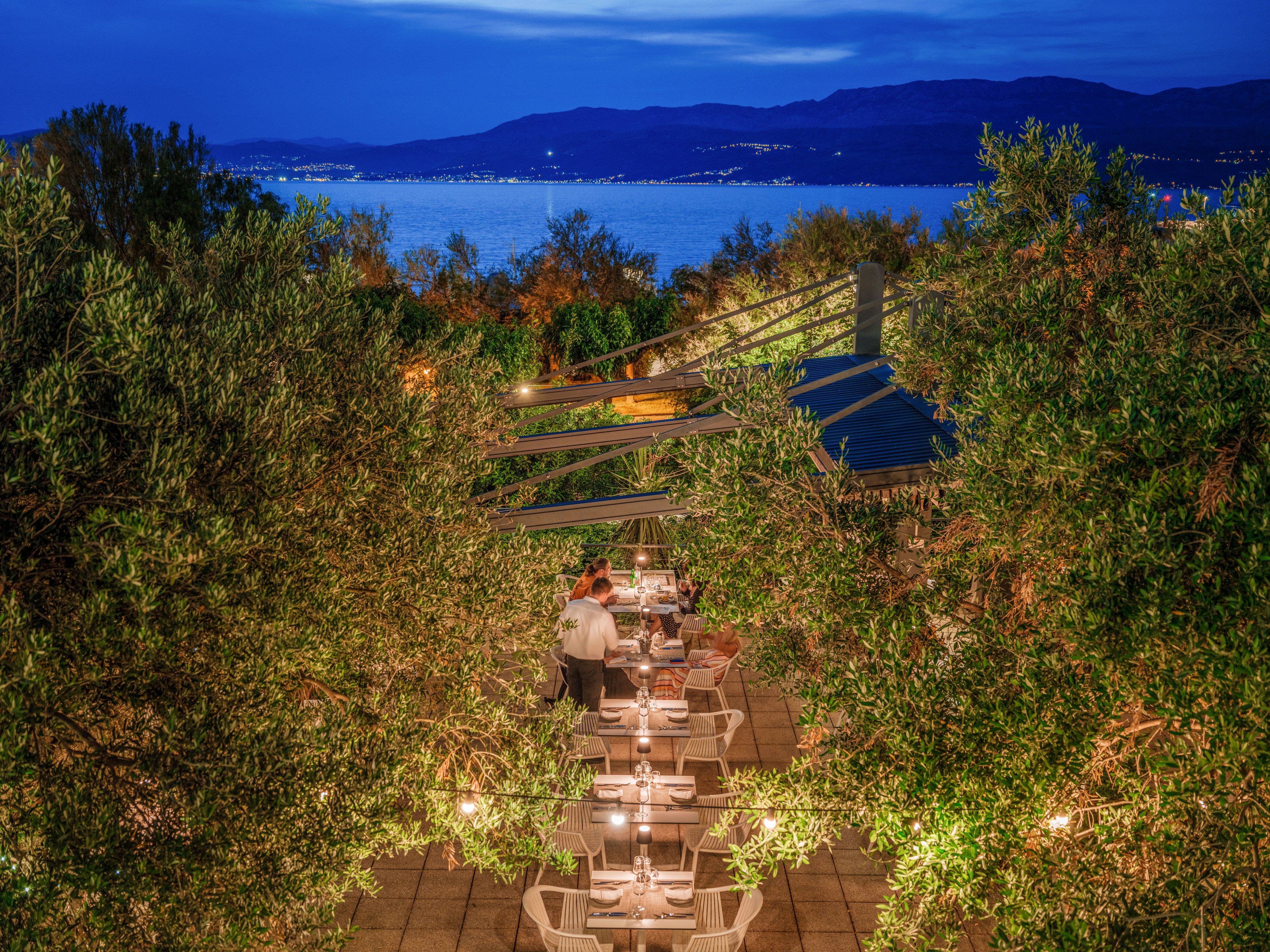 An intimate outdoor restaurant terrace is nestled among lush green trees overlooking a calm sea at dusk. String lights illuminate the dining tables, creating a warm and inviting atmosphere. A server attends to guests seated at white tables, while distant shoreline lights sparkle across the water. The deep blue sky and surrounding foliage frame the cozy evening dining scene.