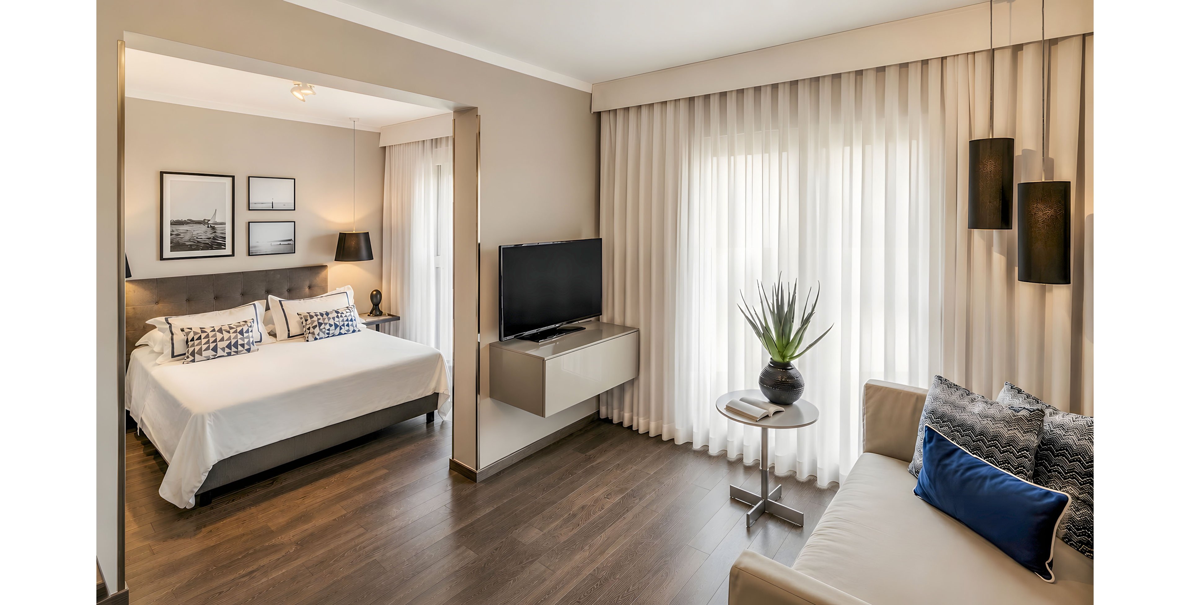 Contemporary hotel-style room featuring a double bed and a cozy sofa seating area. A flat-screen television is mounted on a floating console between the sleeping and living spaces. Large windows with sheer and blackout curtains fill the room with soft natural light. Neutral tones, wooden flooring, framed wall art, and a potted plant on a side table create a calm, elegant atmosphere.