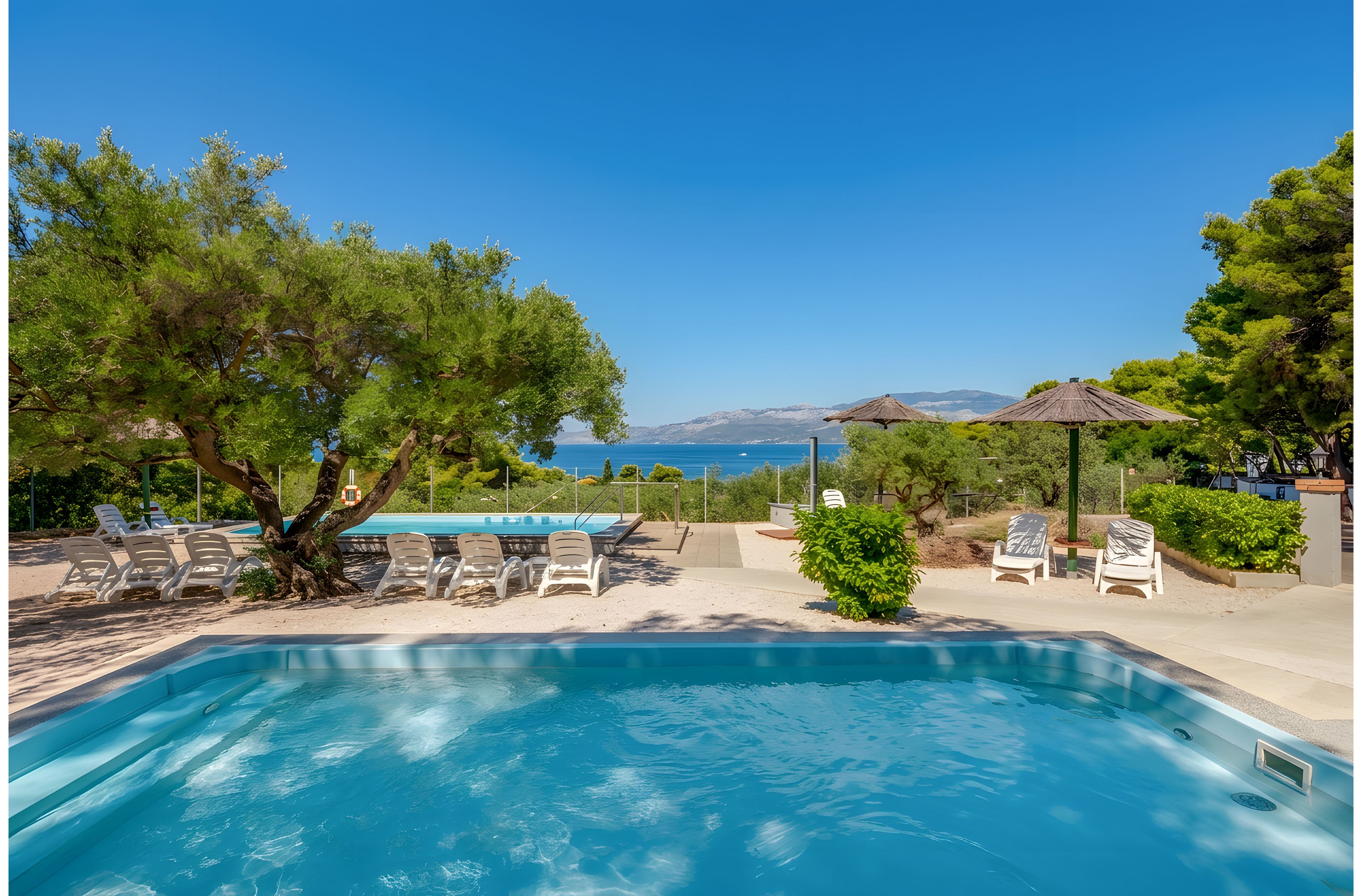 An outdoor swimming pool sits in the foreground of a sunny coastal resort. Lounge chairs and straw parasols are arranged around the poolside terrace, inviting relaxation. Lush green trees frame the scene, with a clear blue sky overhead. In the distance, the calm sea and distant hills create a tranquil vacation atmosphere.