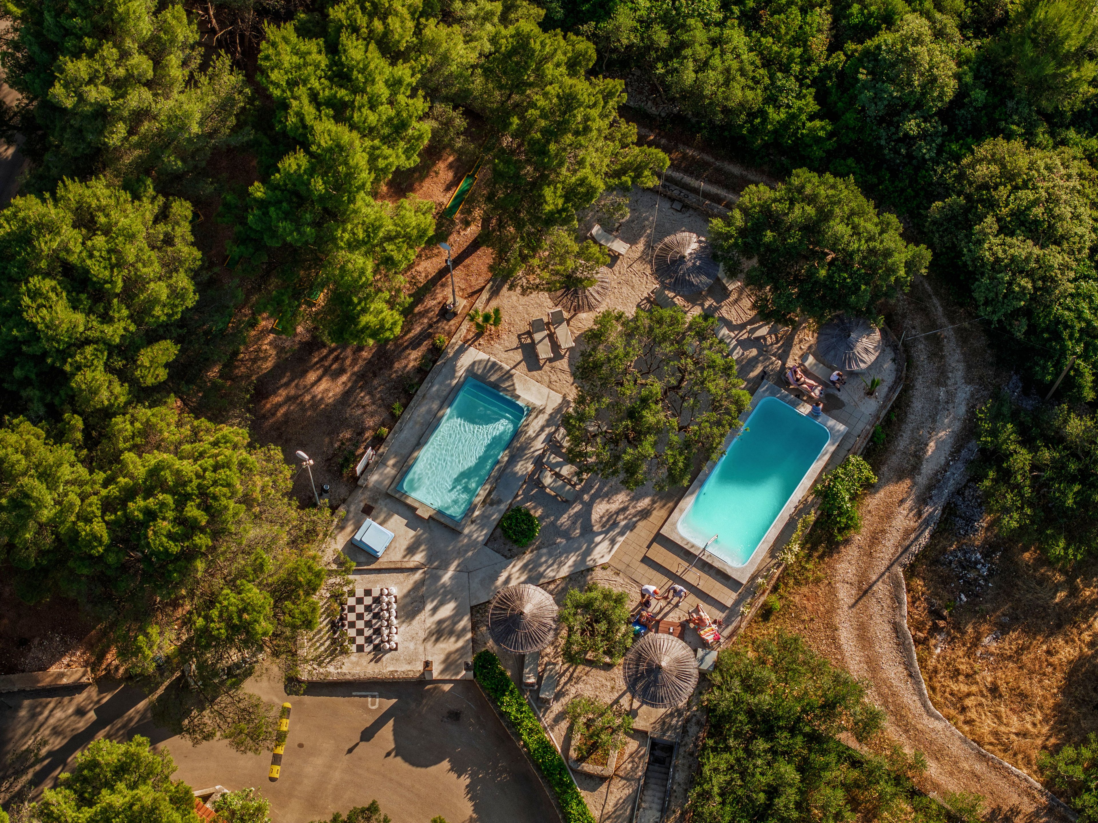 A pair of outdoor swimming pools is nestled within a dense forest setting, viewed from above. One rectangular pool and a smaller irregular-shaped pool are surrounded by sun loungers, umbrellas, and paved walkways. Trees and natural greenery frame the leisure area, with paths and a small gathering of people visible near the pools. The warm lighting suggests a relaxed vacation atmosphere in a secluded resort or holiday retreat.