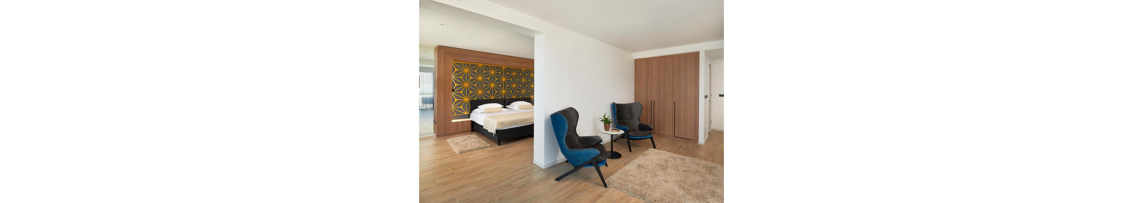 A contemporary hotel suite features a double bed with light bedding and a bold geometric accent wall in yellow and dark gray. The room opens into a small lounge area with two blue armchairs, a round side table, and a potted plant. Wood-look flooring and neutral rugs create a warm, minimalist atmosphere. Built-in wooden closets and clean white walls emphasize the modern, uncluttered design.