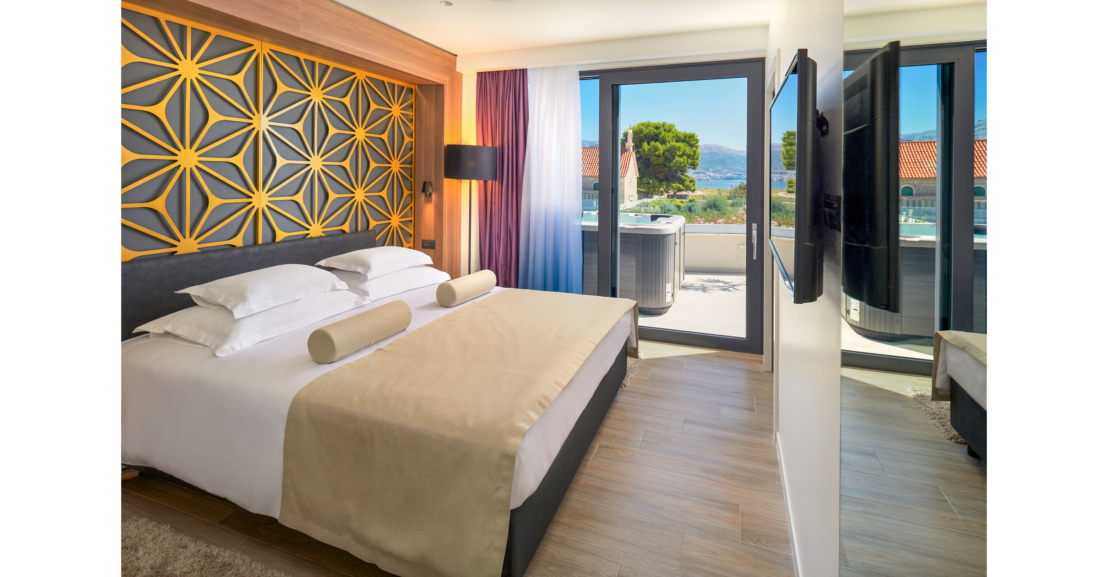 A contemporary hotel bedroom features a large neatly made bed with neutral bedding and cylindrical pillows. The room includes a decorative geometric headboard in yellow and gray tones and a wall-mounted flat-screen television opposite the bed. Floor-to-ceiling glass doors open onto a balcony with outdoor seating and views of rooftops, trees, and distant mountains. Soft natural light and layered curtains create a bright yet cozy atmosphere.