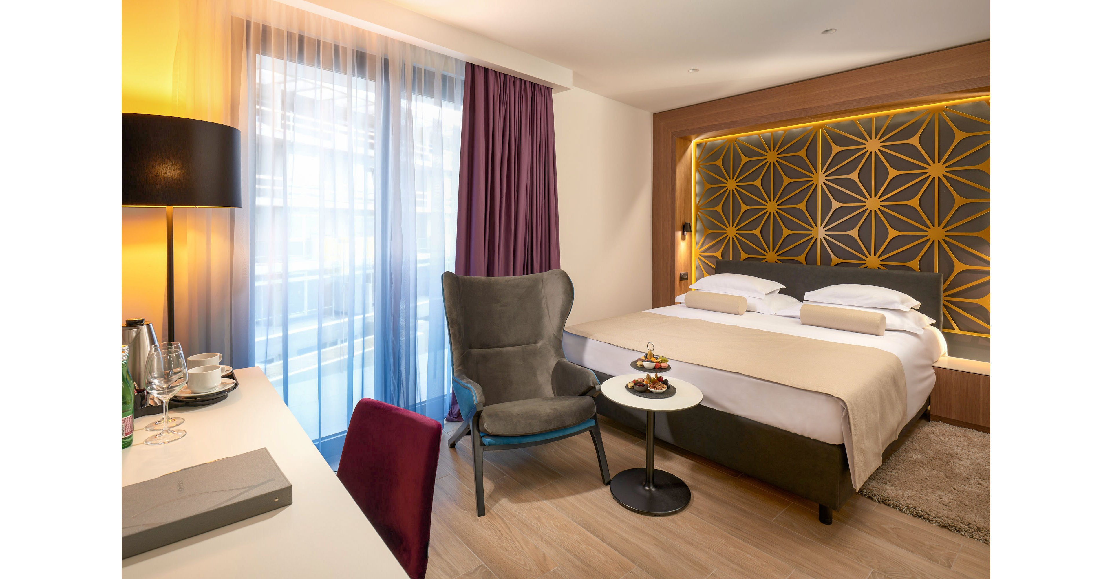A contemporary hotel room features a neatly made double bed with soft pillows and a geometric illuminated headboard. Beside the bed, a small round table holds a dessert tray next to a plush gray armchair. Floor-to-ceiling curtains in white and purple frame the window, allowing warm natural light to mix with ambient interior lighting. A work desk with a chair, kettle, and cups completes the comfortable and functional guest space.