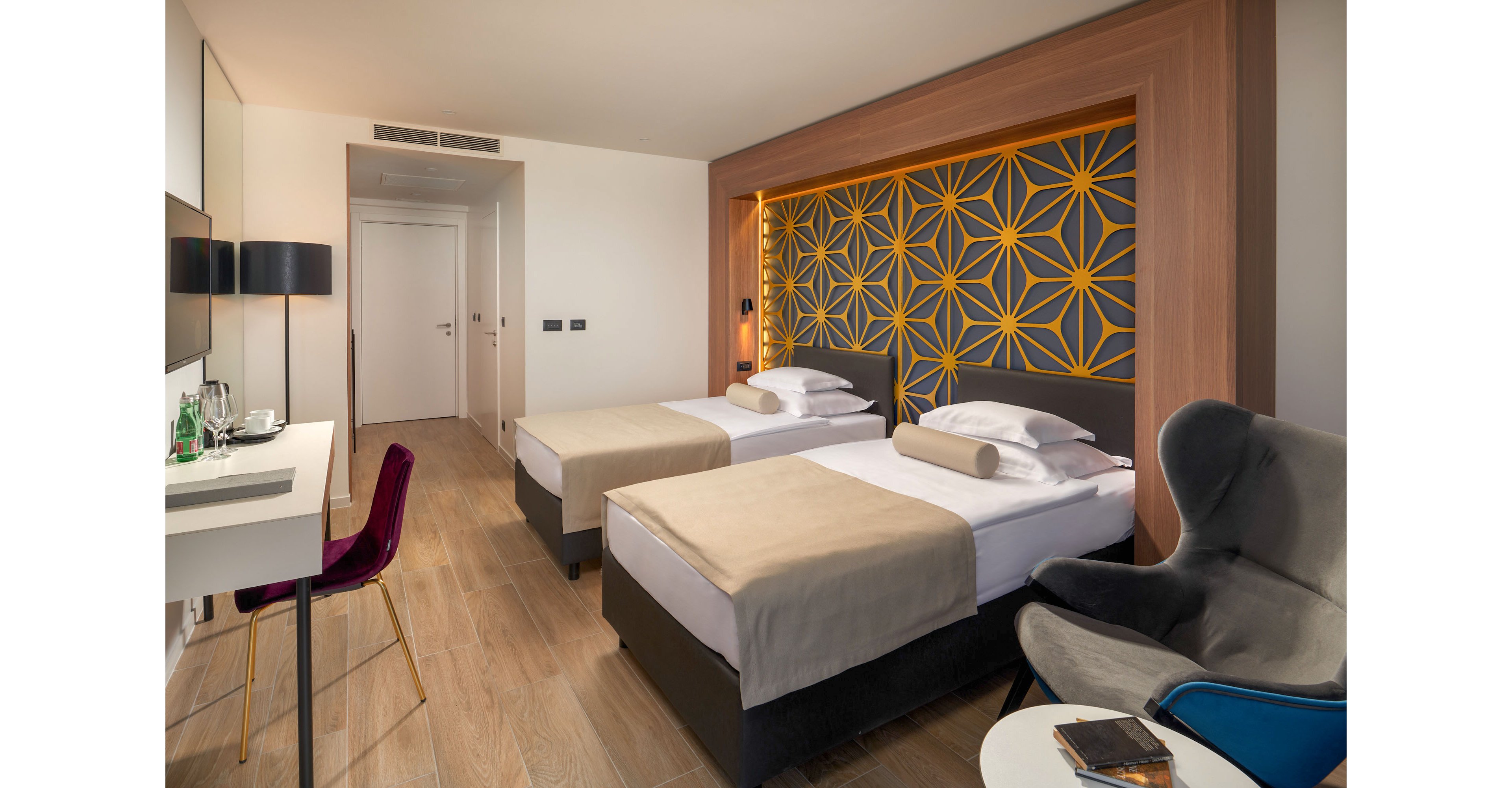 A contemporary hotel room features two neatly made twin beds with beige bedding and white pillows. The headboard wall showcases a bold geometric pattern in yellow and gray, adding a decorative focal point. A desk with a chair, bottled water, and glasses sits opposite the beds, while a plush armchair and small table create a cozy seating area. The space is bright, minimalist, and designed for comfortable short stays.