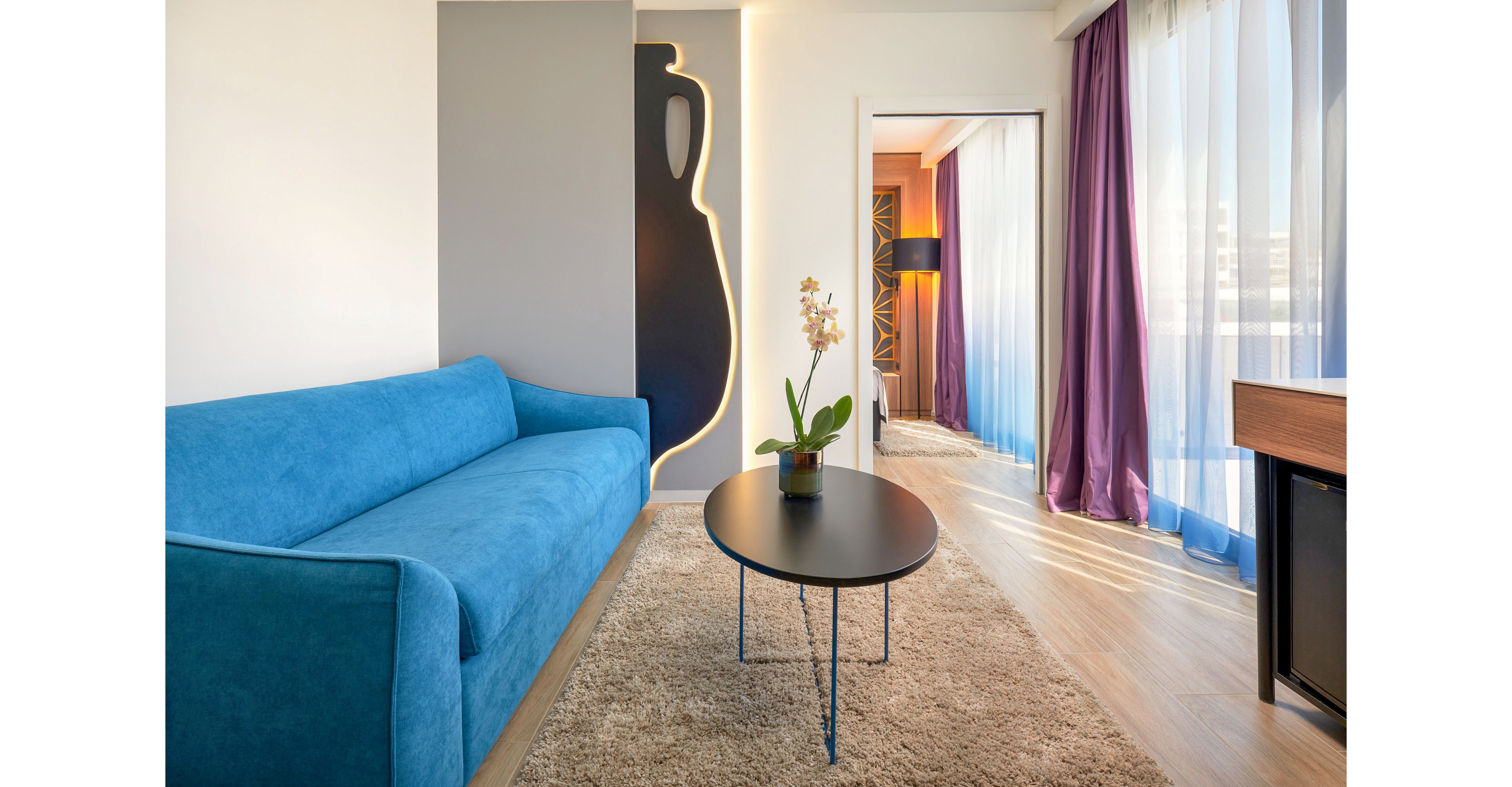 A contemporary hotel living area features a bright blue sofa set against a decorative wall with a vase-shaped cutout and soft backlighting. A round black coffee table with thin metal legs stands on a beige rug, holding a potted orchid. Floor-to-ceiling windows with sheer white and purple curtains allow natural light to fill the room. In the background, a doorway leads to another space with a standing lamp and wooden accents, enhancing the modern, cozy atmosphere.