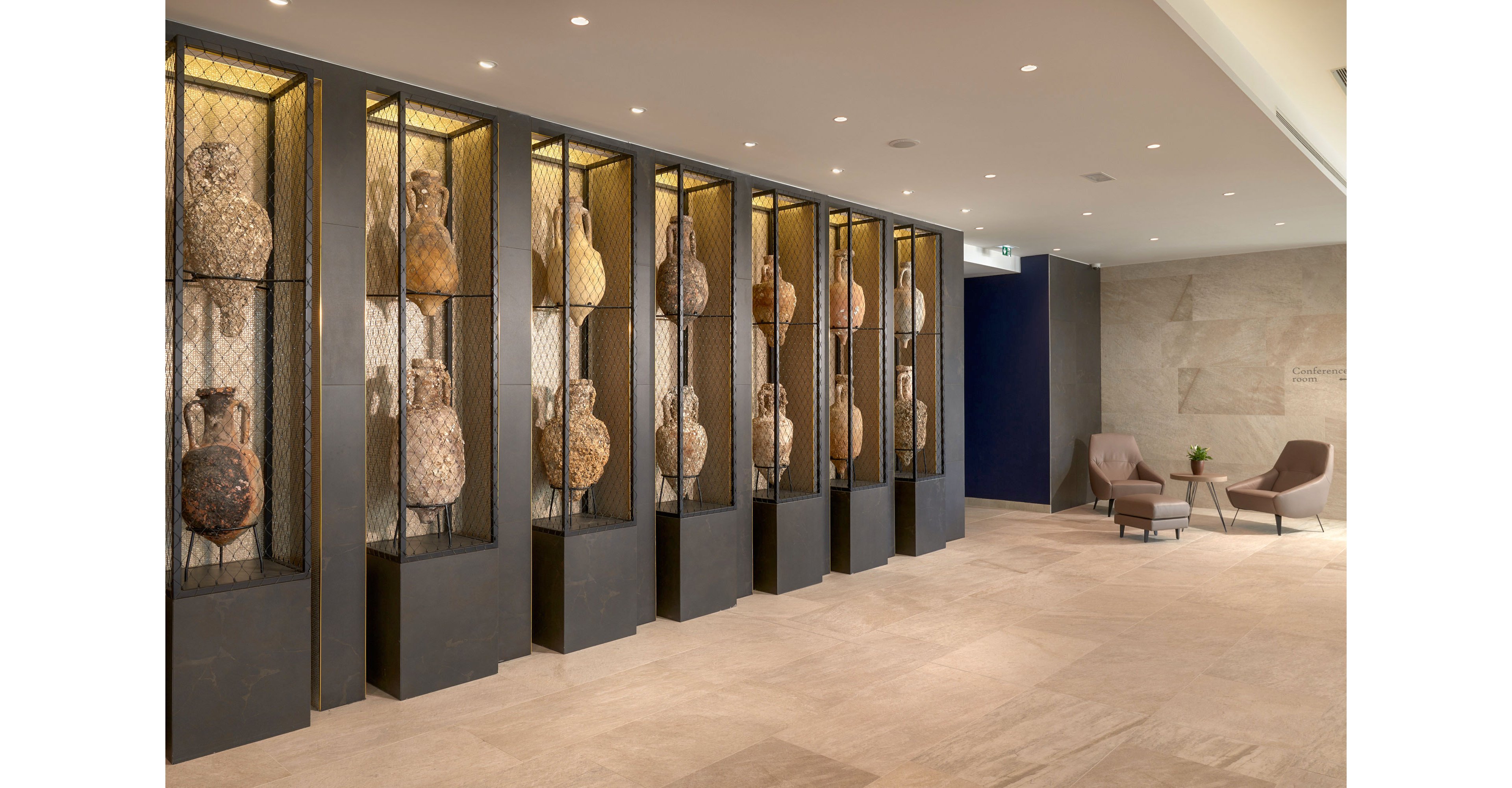 A contemporary lobby features a long row of tall display cases holding large, weathered pottery vessels. The amphora-like artifacts are arranged vertically behind mesh panels, creating a museum-style presentation along one wall. On the opposite side, minimalist lounge chairs and a small table provide a quiet seating area. Neutral beige flooring and walls, combined with warm recessed lighting, give the space a calm and sophisticated atmosphere.