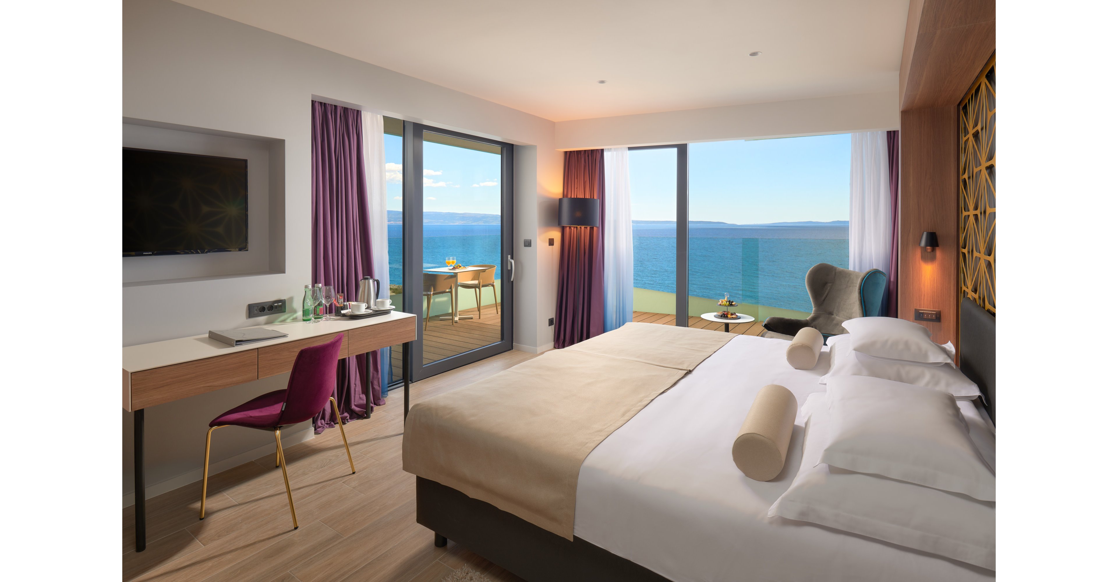 A contemporary hotel room features a large neatly made bed facing floor-to-ceiling windows. The space includes a desk with a flat-screen TV, chair, and a tray with water and glasses. Sliding glass doors open to a balcony with seating and a panoramic ocean view. Soft neutral tones are accented by purple curtains and modern decor, creating a calm and inviting atmosphere.