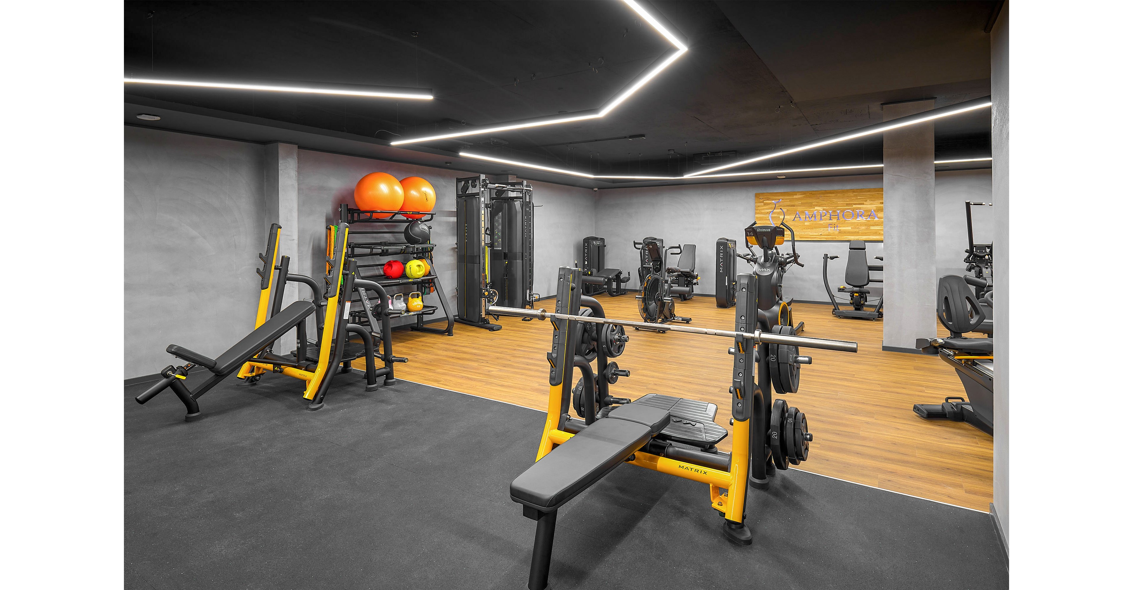 A contemporary indoor gym features various strength training machines and free weights. In the foreground, a yellow weight bench with a loaded barbell stands on a dark rubber floor. Additional equipment such as squat racks, cable machines, and exercise balls are arranged neatly around the room. The space has a minimalist design with concrete walls, wooden flooring sections, and linear LED ceiling lights creating a sleek atmosphere.