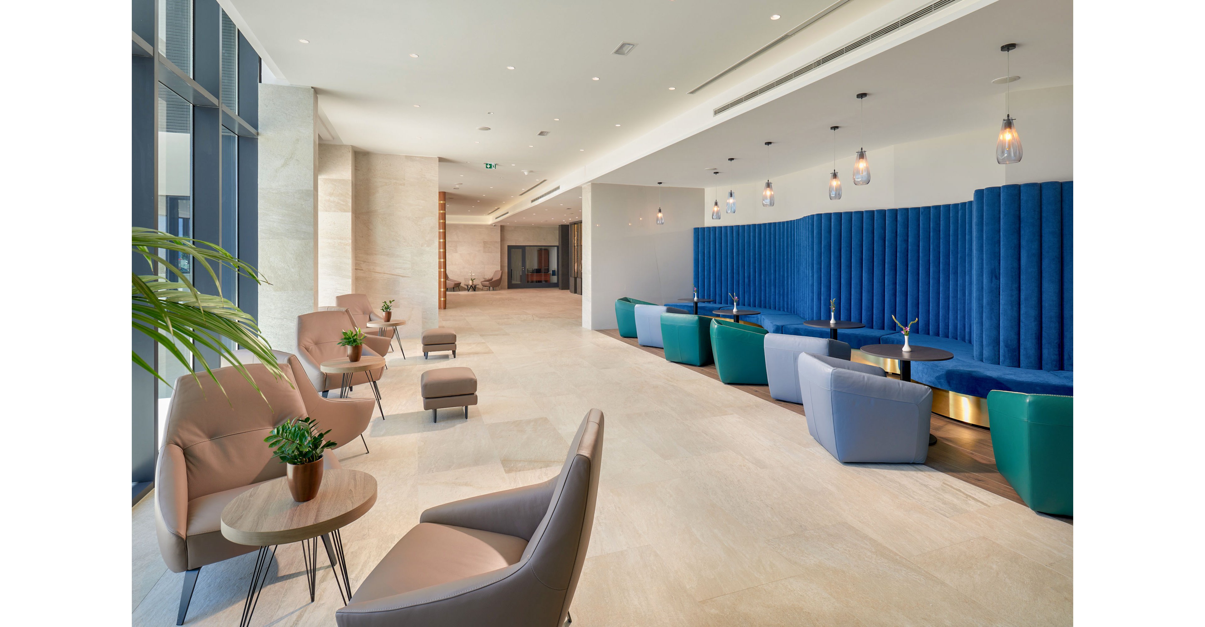 A contemporary lounge area features a mix of armchairs and low tables arranged along a bright corridor. On the right side, a long blue upholstered banquette with small round tables creates a cozy seating zone. Pendant lights hang from the ceiling, casting warm light over the neutral stone floor. Large windows on the left bring in natural daylight and reveal a glimpse of greenery, enhancing the airy, welcoming atmosphere.