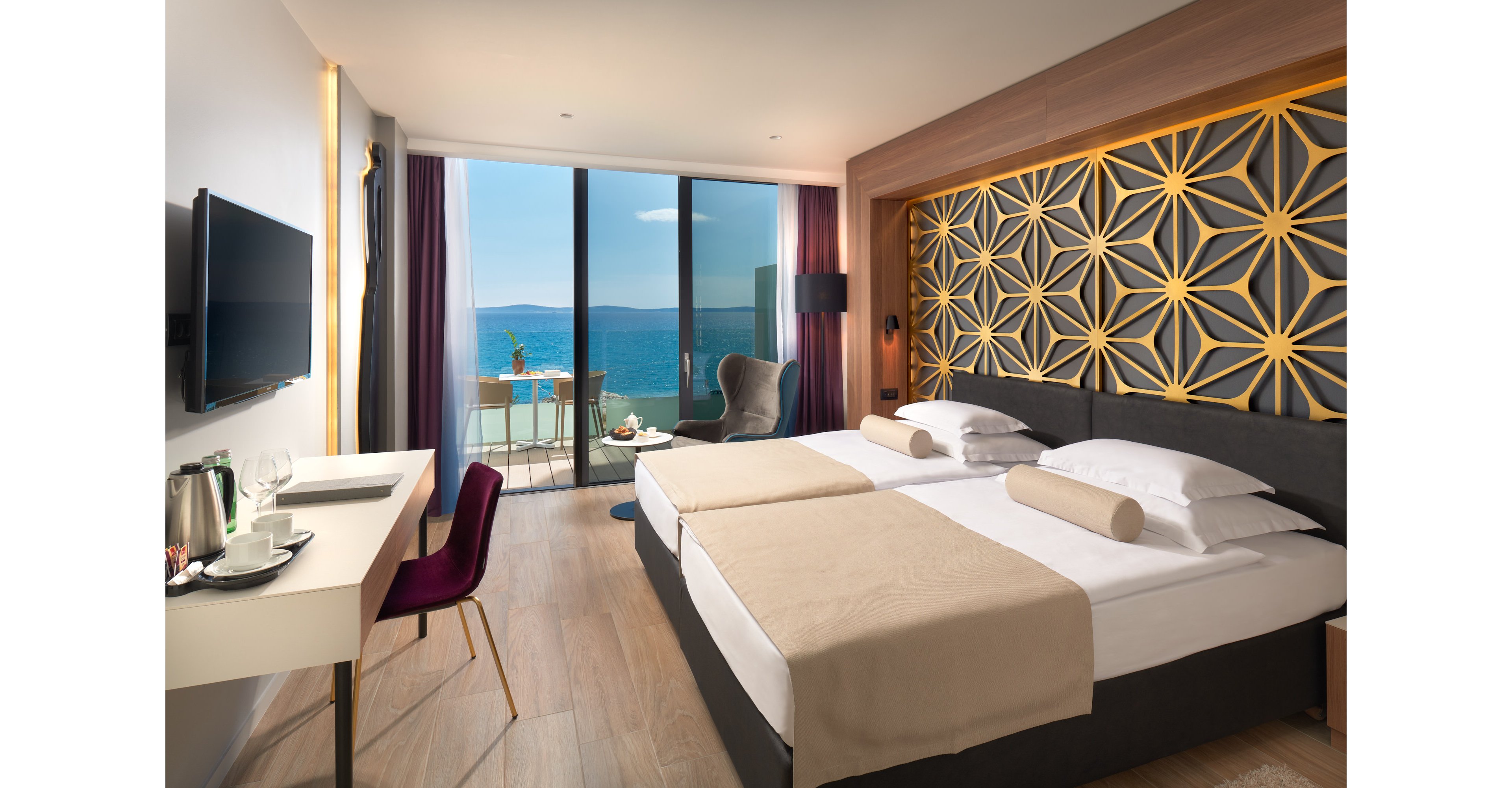 A contemporary hotel room features two neatly made twin beds with beige throws and cylindrical pillows. The space includes a desk with a television, chair, and a tray with a kettle and cups. Large floor-to-ceiling glass doors open onto a balcony with chairs and a table overlooking a bright blue ocean. Decorative geometric wall panels and warm wood flooring create a stylish, relaxing atmosphere.
