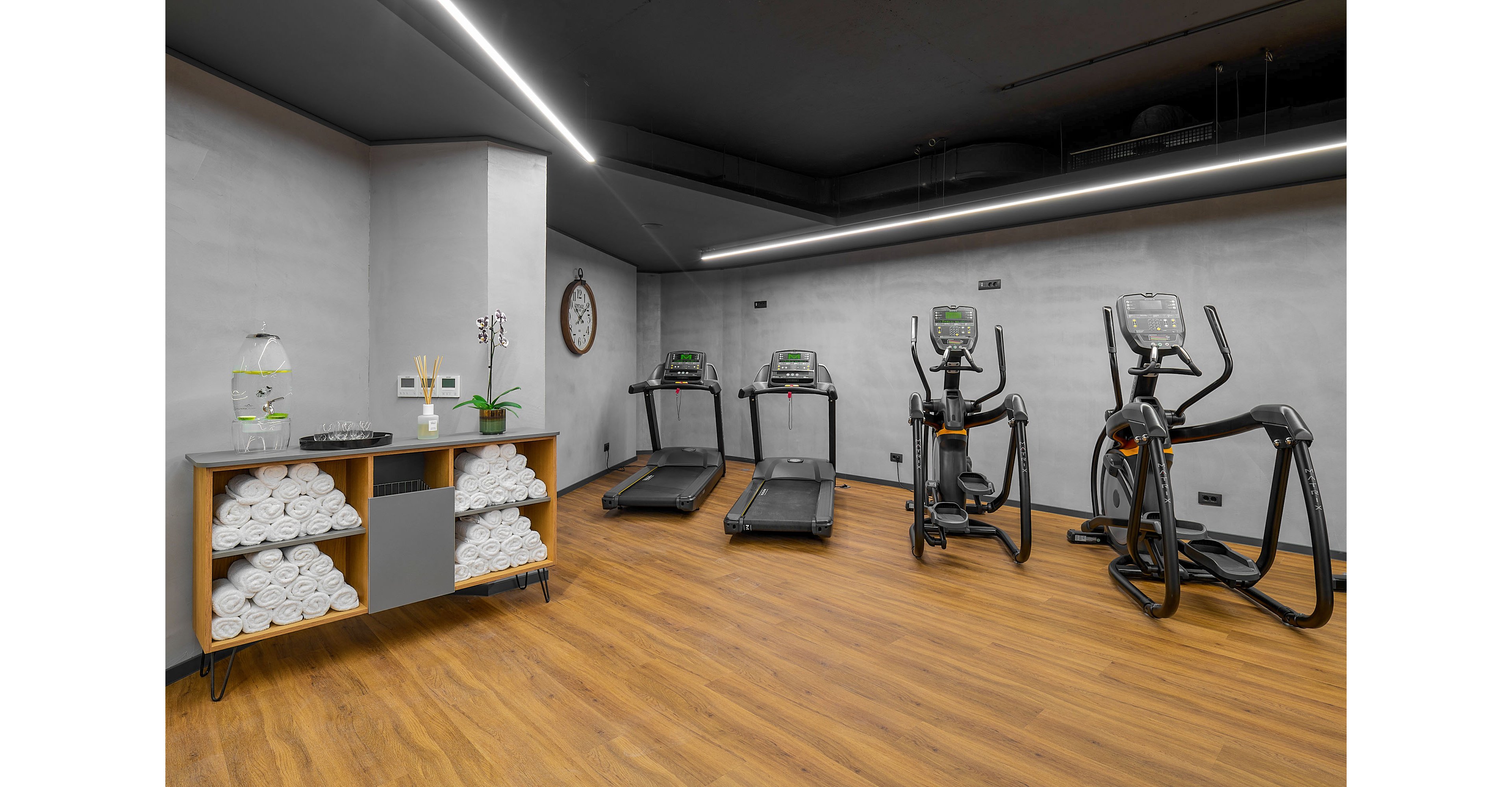 A contemporary indoor gym features several treadmills and elliptical machines arranged along a wall. The room has a clean, minimalist design with gray walls, a dark ceiling, and warm wooden flooring. On the left, a shelving unit is neatly stocked with rolled white towels, water dispensers, and small decor items. A wall clock and linear ceiling lights add functional details to the bright, organized space.