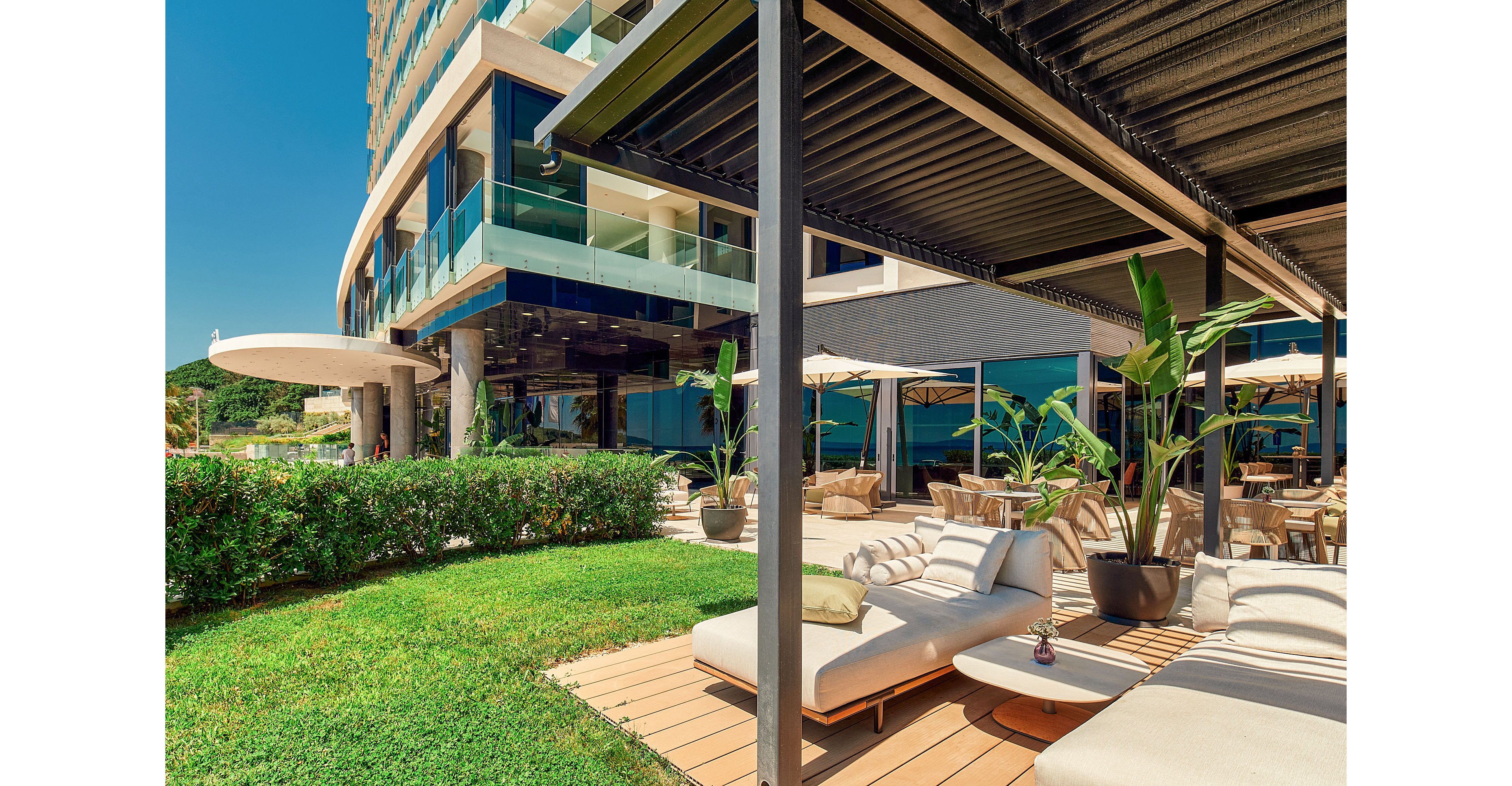 A contemporary hotel terrace features cushioned sun loungers arranged on a wooden deck. The outdoor area is shaded by a sleek pergola and surrounded by lush green grass and manicured hedges. Large glass windows of the building reflect the bright blue sky and nearby landscape. Umbrellas, potted plants, and outdoor seating create a relaxed resort atmosphere.