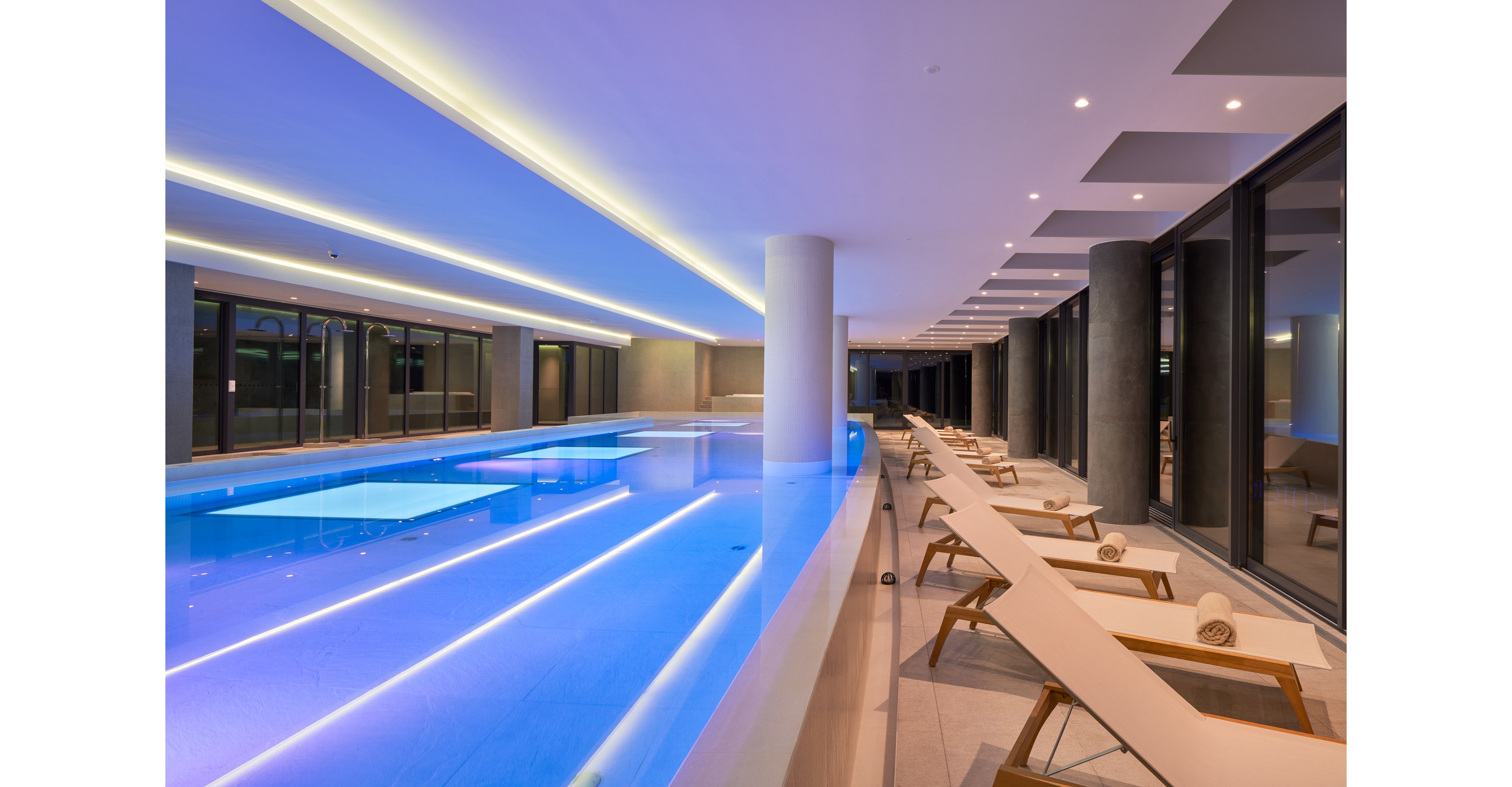 A sleek indoor swimming pool is illuminated with soft blue lighting in a contemporary building. Long rows of cushioned lounge chairs line one side of the pool, each with neatly rolled towels. Floor-to-ceiling windows reflect the water and reveal the dark exterior outside. Recessed ceiling lights and clean architectural lines create a calm, luxurious spa-like atmosphere.