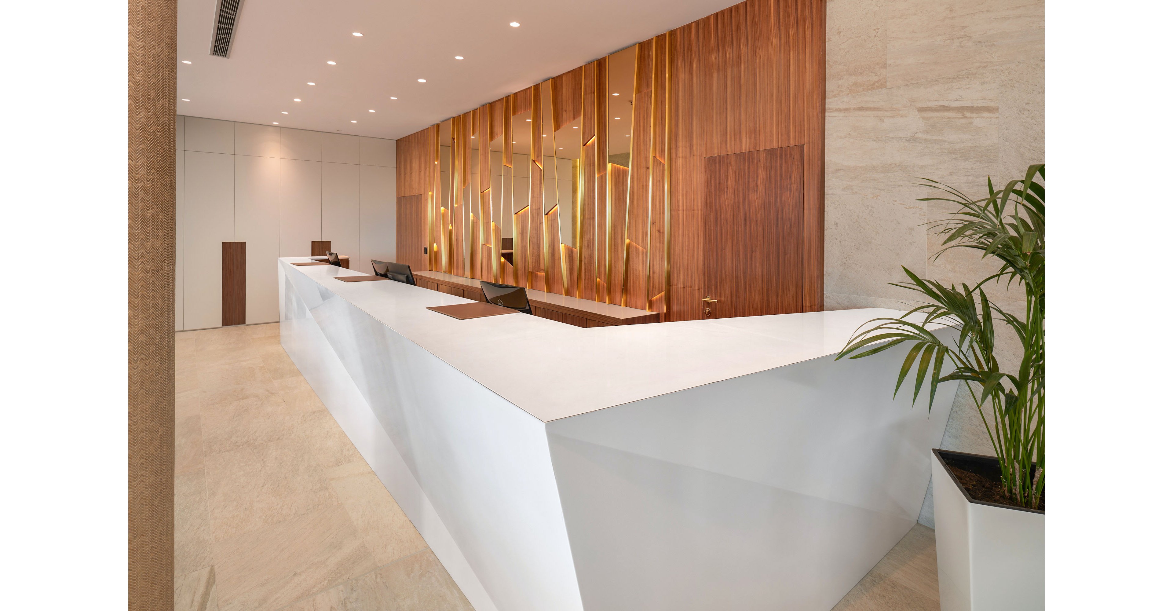 A sleek white geometric reception desk dominates a contemporary office lobby. Warm wooden wall panels with vertical golden accents create a sophisticated backdrop behind the counter. Recessed ceiling lights evenly illuminate the clean, minimalist space. A potted green plant adds a touch of nature and contrast to the neutral interior palette.