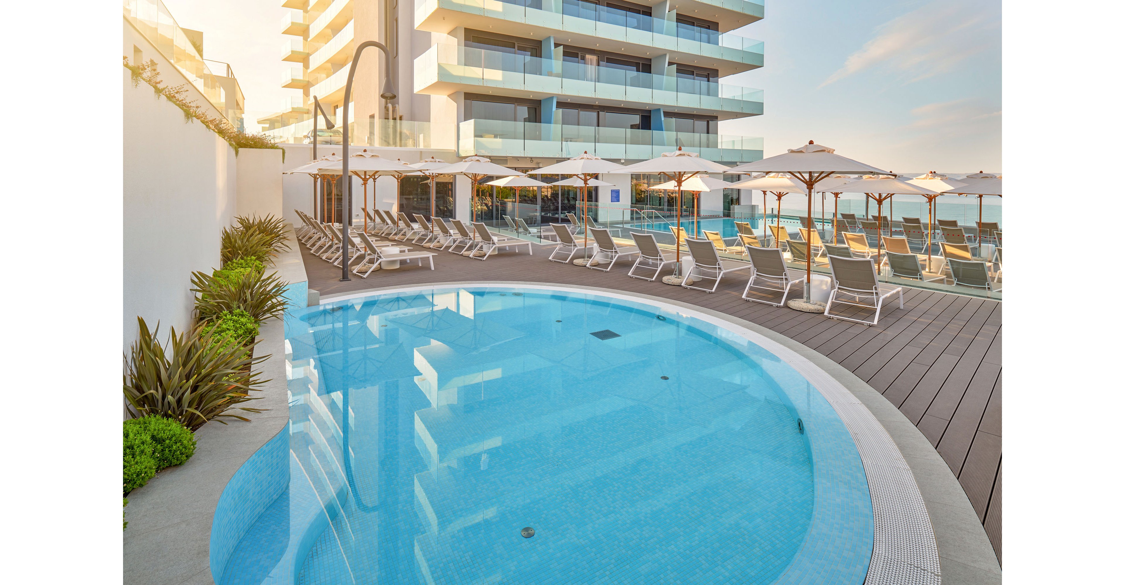 A curved outdoor swimming pool sits beside a modern hotel or apartment building with glass balconies. Numerous sun loungers and white parasols line the wooden deck, creating a resort-like atmosphere. The scene is bathed in warm early morning or late afternoon light, with clear blue water reflecting the sky. Decorative plants soften the edges of the pool area, suggesting a relaxing vacation setting.