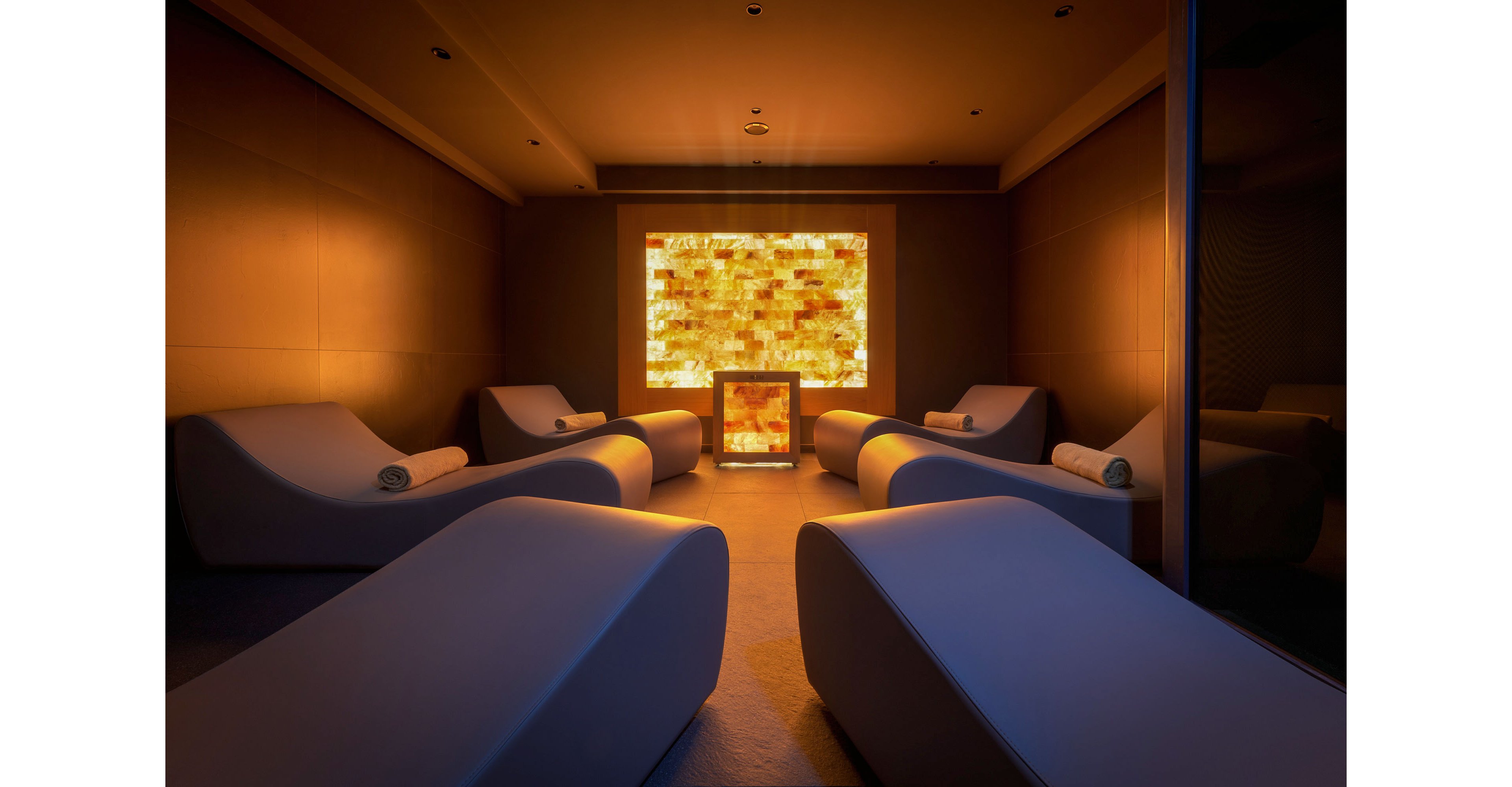 A dimly lit spa relaxation room features several modern curved loungers arranged symmetrically. Each lounger has a rolled towel placed at the headrest, emphasizing comfort and readiness for guests. A glowing illuminated wall panel at the far end creates a warm amber focal point in the otherwise dark space. The overall atmosphere is tranquil and luxurious, ideal for rest and meditation.
