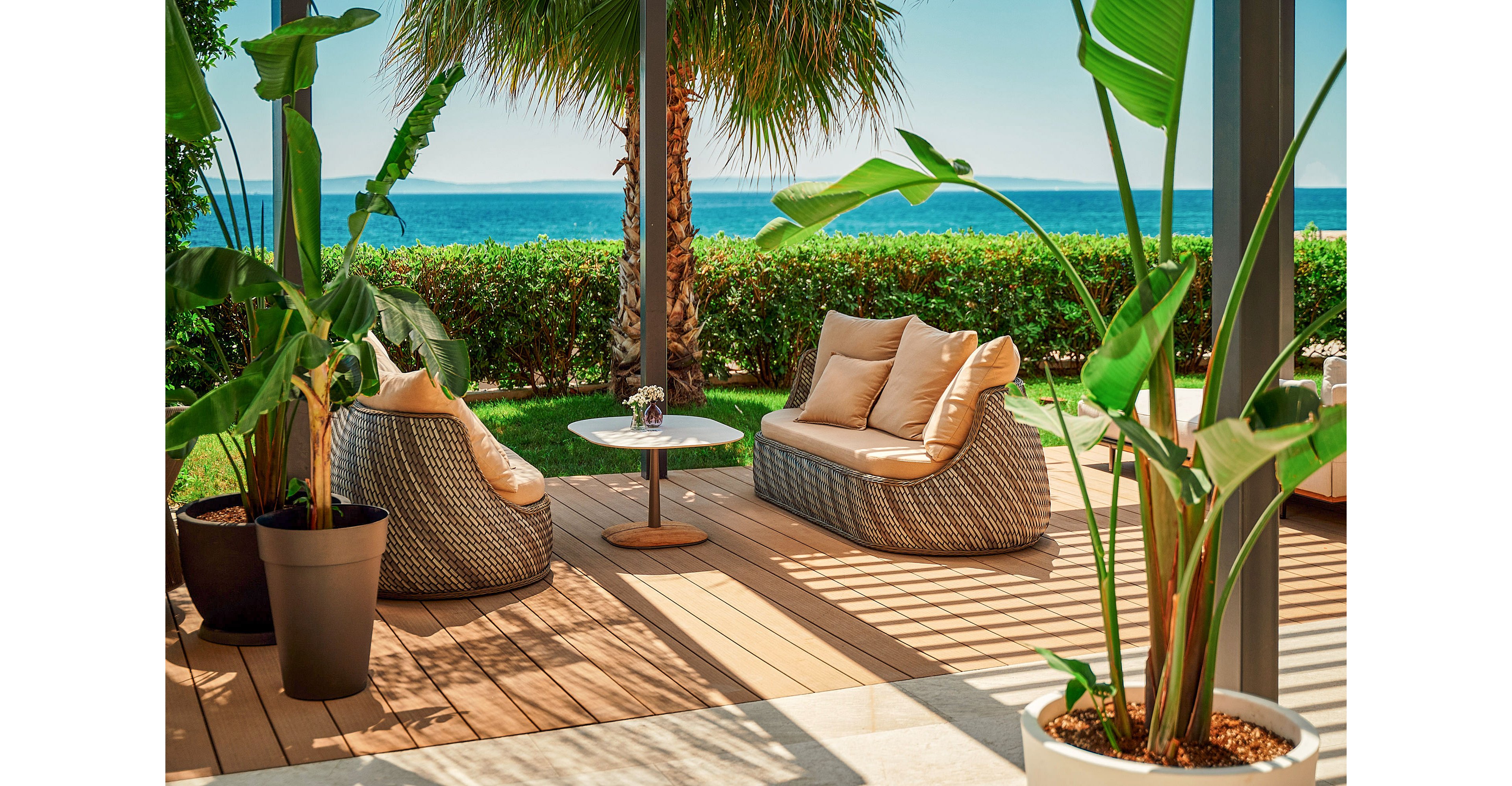 An inviting outdoor lounge area features two modern wicker sofas with light cushions arranged around a small round table. The patio is shaded by a pergola and surrounded by lush green plants in large pots. A tall palm tree and manicured hedge frame the view of the calm blue sea and distant horizon. Warm sunlight casts soft shadows across the wooden deck, creating a relaxed coastal atmosphere.