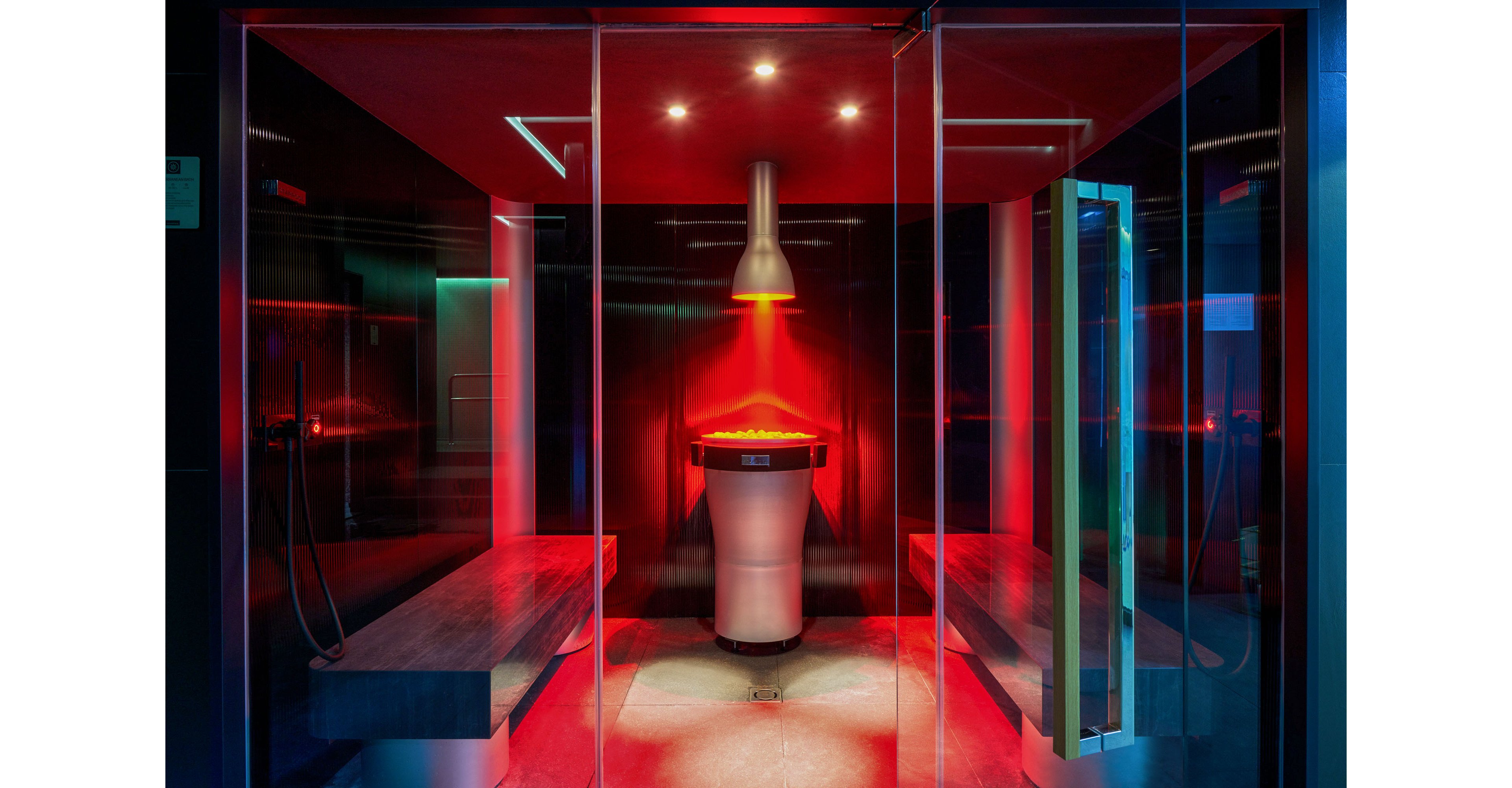 A contemporary steam room interior is illuminated with intense red lighting focused on a central column feature. The space is enclosed by glass walls and doors, creating a sleek, transparent boundary. Long stone benches line both sides of the room, suggesting a spa or wellness environment. Overhead spotlights and the glowing central fixture add a futuristic, atmospheric feel to the scene.