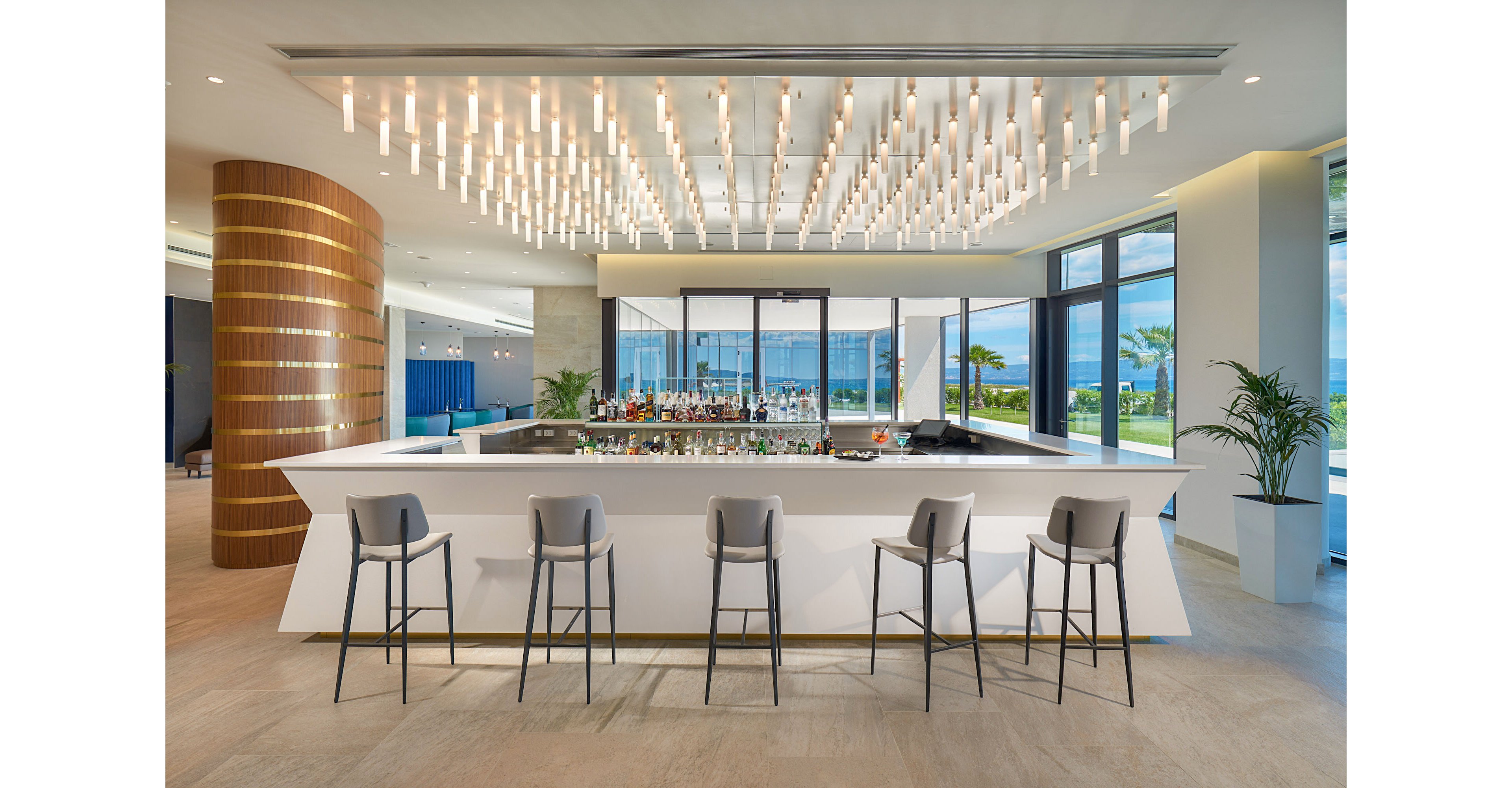 A contemporary hotel bar features a sleek white counter surrounded by minimalist gray bar stools. The space is illuminated by a dramatic ceiling installation of hanging lights, creating a bright and inviting atmosphere. Floor-to-ceiling windows reveal a view of the ocean, palm trees, and manicured lawns outside. Warm wood accents and neutral tones contribute to a clean, upscale, and modern interior design.