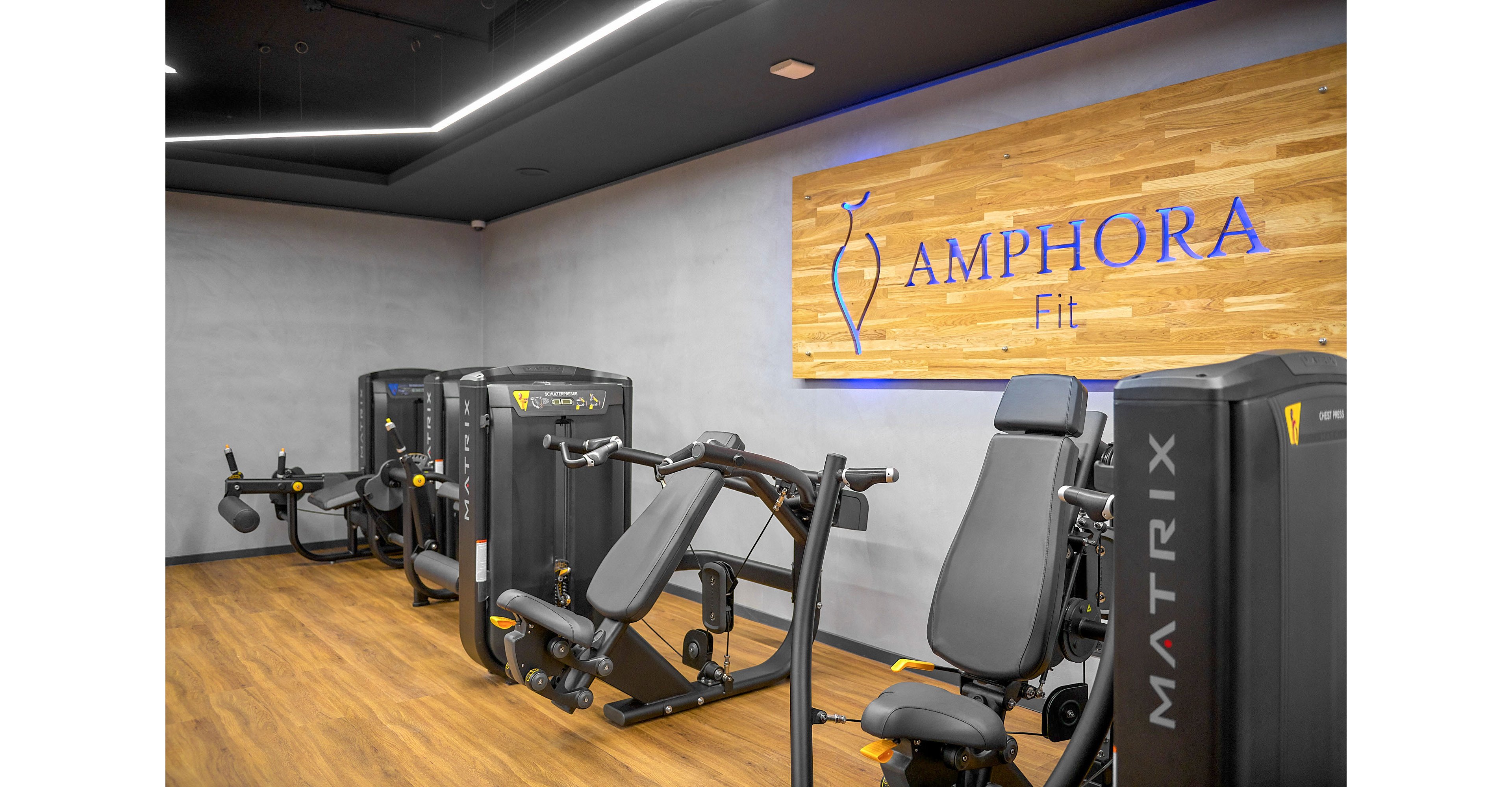 A modern fitness studio features several strength training machines neatly arranged along a wall. The room has a clean design with light gray walls, wood-style flooring, and dark ceiling panels with linear lighting. A large wooden wall panel displays the text "AMPHORA Fit" with a stylized amphora icon. The exercise equipment is labeled with the word "MATRIX" on the side of one machine.