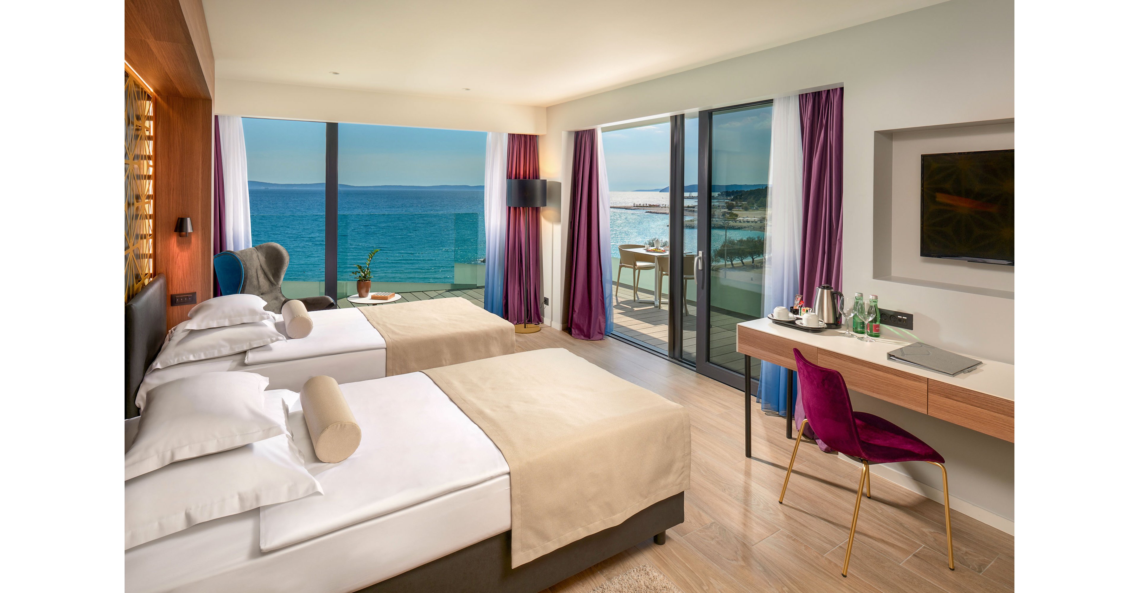 A contemporary hotel room features two neatly made twin beds facing a large window. Floor-to-ceiling glass doors open onto a balcony overlooking a bright blue sea and distant coastline. A sleek desk with a magenta chair, bottled water, and a kettle sits opposite a wall-mounted television. Soft neutral bedding, wooden flooring, and colorful curtains create a calm, inviting atmosphere filled with natural light.