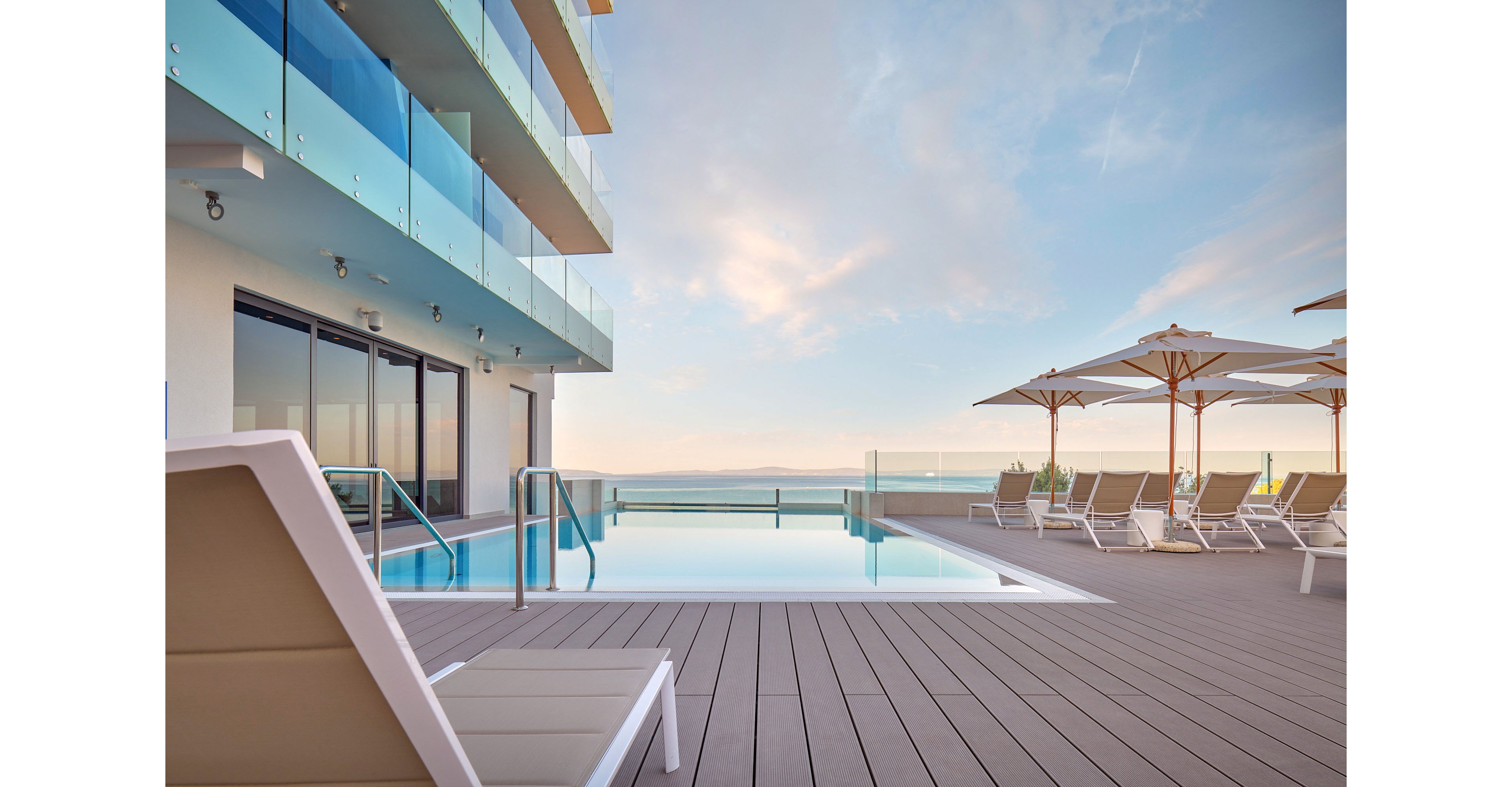 A modern rooftop swimming pool sits beside a contemporary building with glass balconies. Lounge chairs and large umbrellas line the wooden deck, facing the calm water. The scene overlooks a distant horizon and sea under a soft pastel sky. Warm early morning or late afternoon light creates a tranquil, resort-like atmosphere.