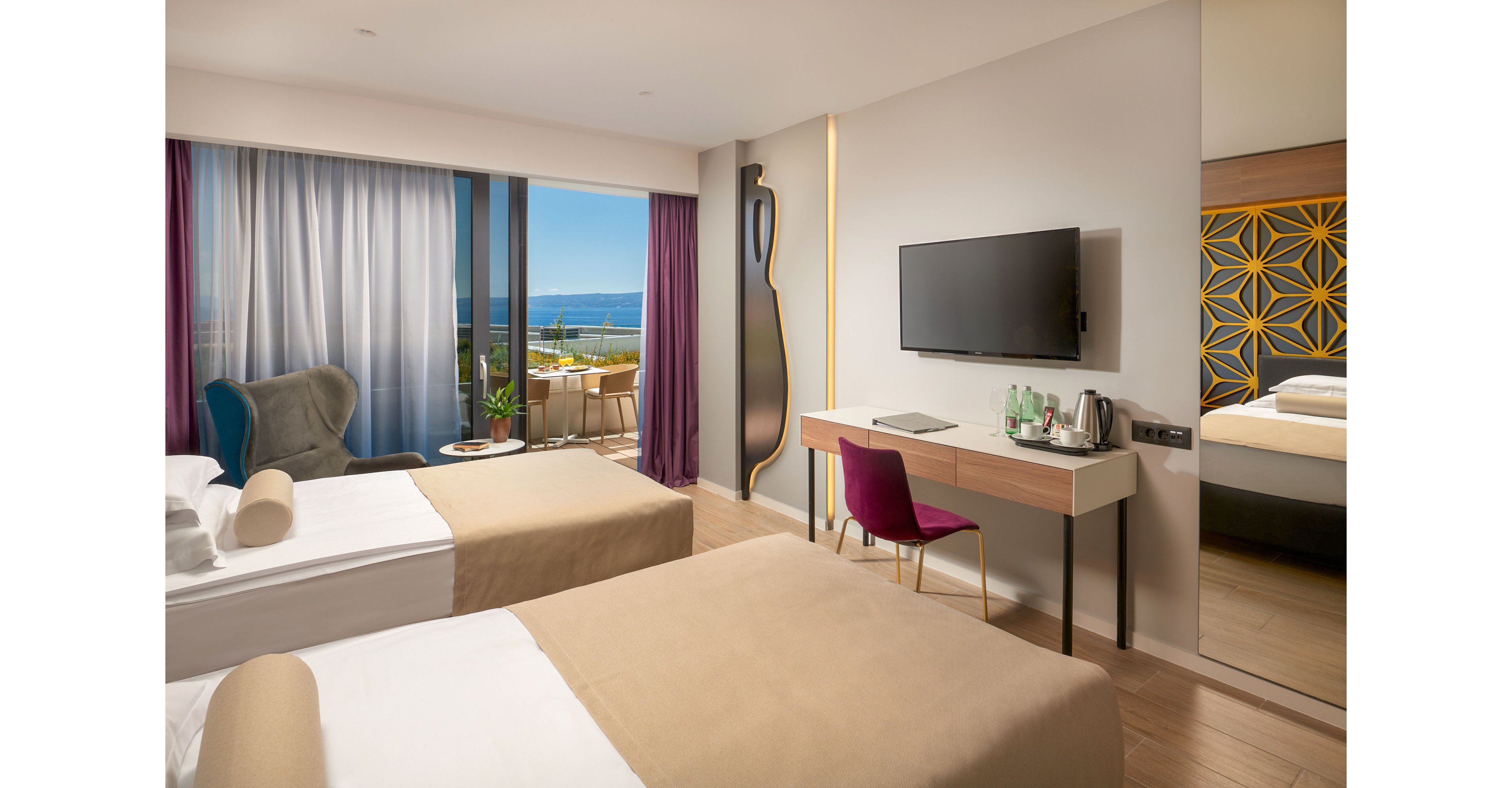 A contemporary hotel room features two neatly made twin beds with beige bedding and white pillows. A large sliding glass door opens to a balcony overlooking the sea and distant coastline. The interior includes a wall-mounted flat-screen TV, a minimalist desk with a magenta chair, and a full-length mirror reflecting geometric wall art. Soft neutral tones, accent lighting, and floor-to-ceiling curtains create a calm, inviting atmosphere.