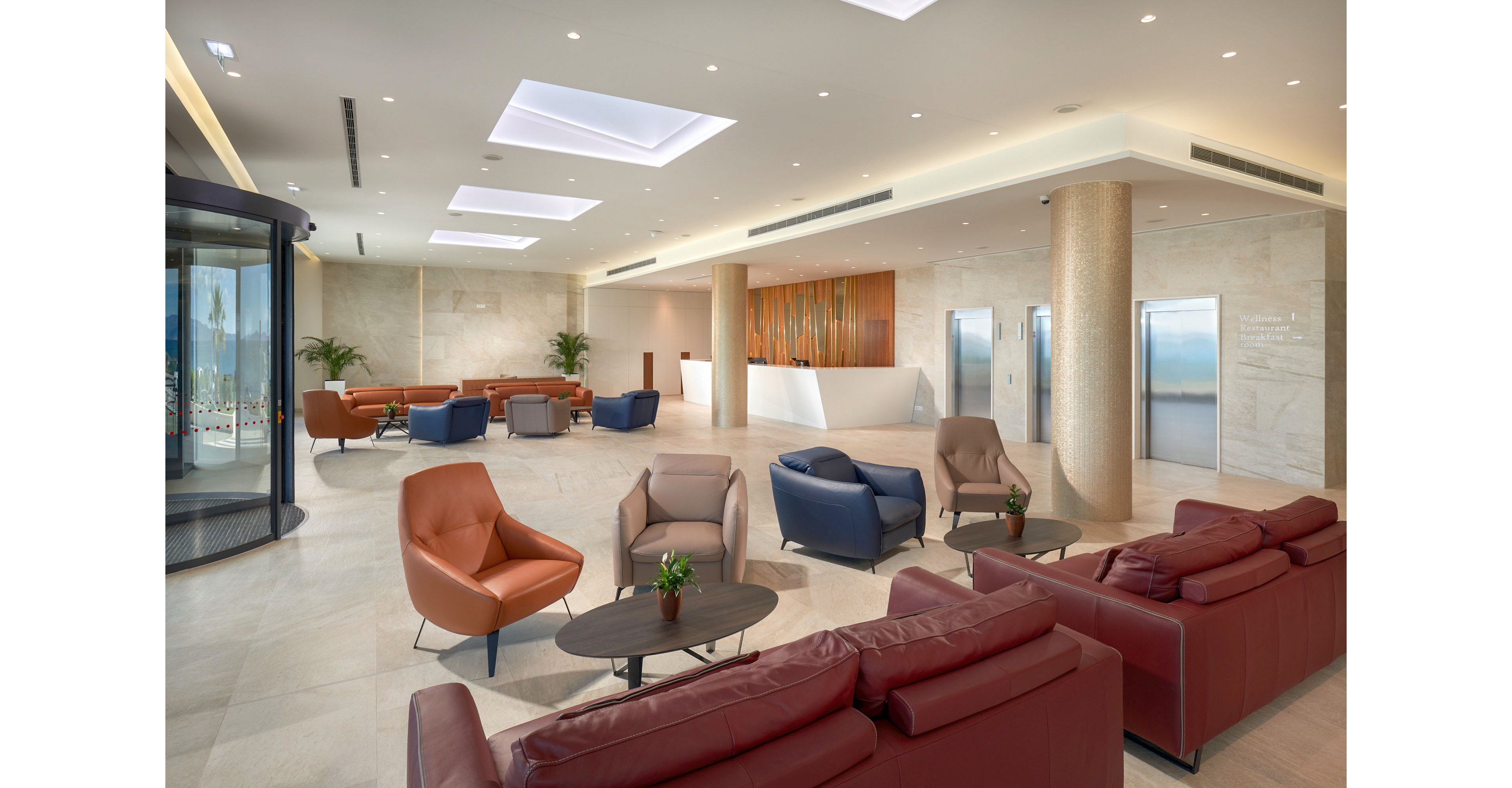 A spacious modern lobby features a white reception desk and polished stone floors. Multiple seating areas with leather armchairs and sofas are arranged around low coffee tables with small plants. Recessed ceiling lights and skylights create bright, even illumination throughout the space. Elevators and a glass revolving door are visible in the background, emphasizing a contemporary corporate environment.