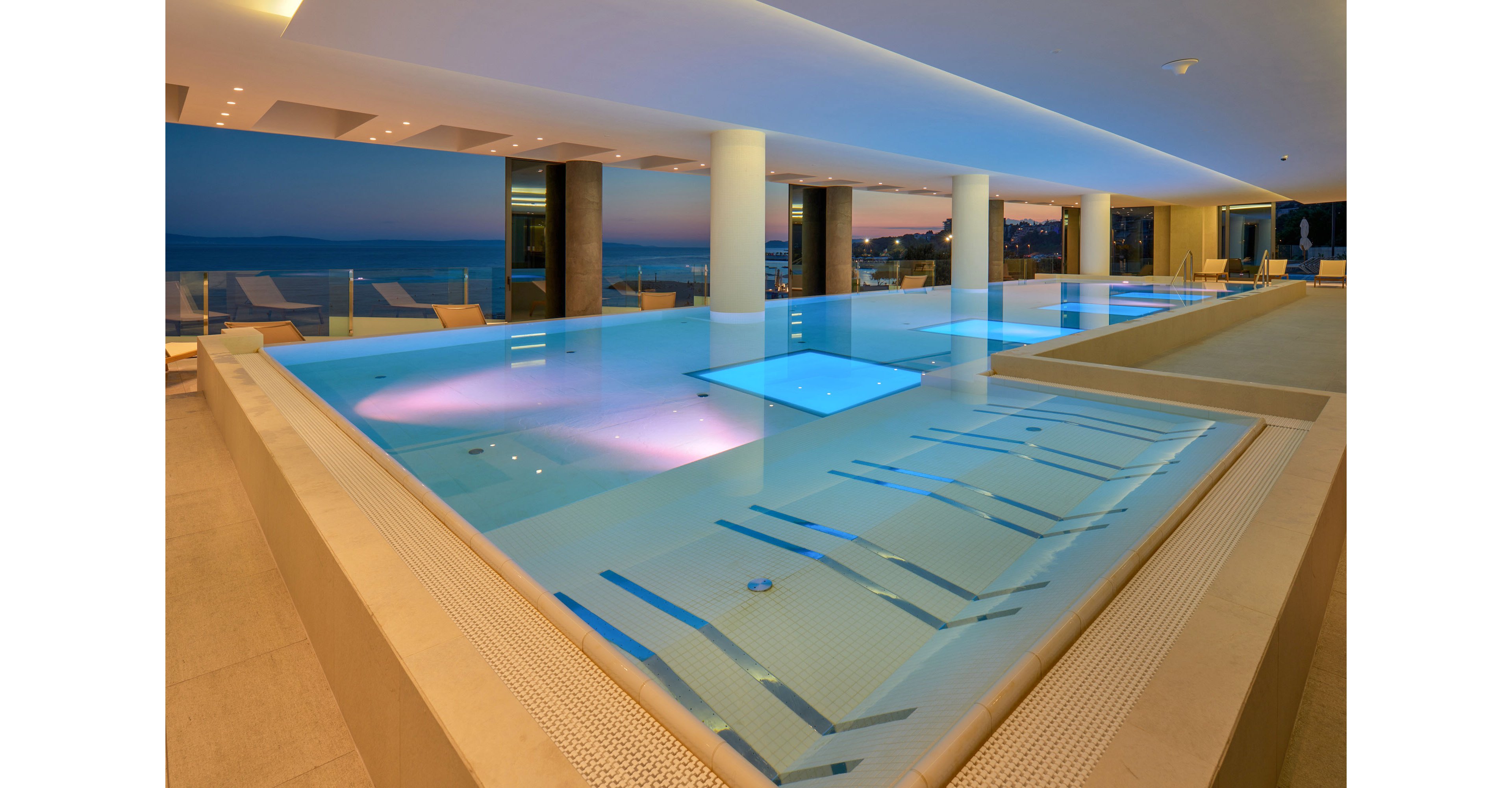 A contemporary indoor pool area features multiple interconnected pools with built-in loungers and underwater lighting. Soft blue and pink lights illuminate the water, creating a luxurious spa-like atmosphere. Large windows open to a coastal view at dusk, with the sea and shoreline visible in the distance. Minimalist lounge chairs line the terrace, enhancing the serene and upscale resort setting.
