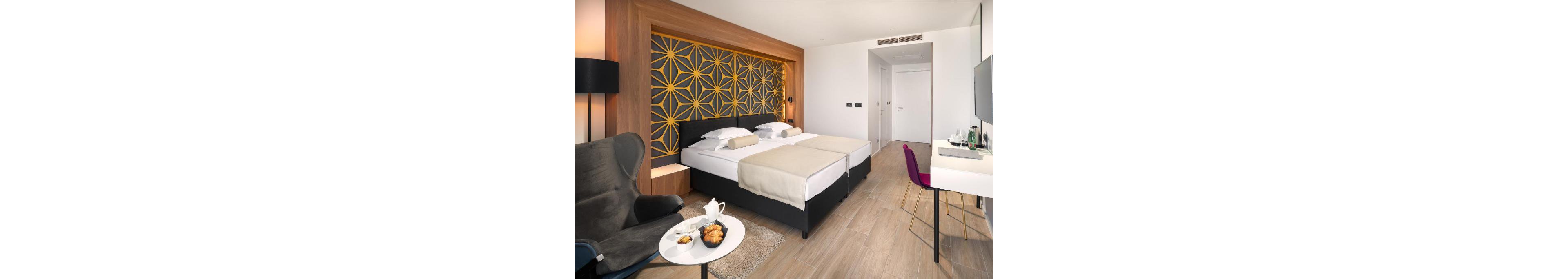 A contemporary hotel room features two neatly made twin beds with light beige throws and white linens. The headboard wall showcases a bold geometric pattern in yellow and dark gray framed by warm wood paneling. A cozy seating area with an armchair and small round table holds a teapot and pastries, while a sleek desk with a magenta chair lines the opposite wall. Neutral tones, clean lines, and soft lighting create a calm, inviting atmosphere.