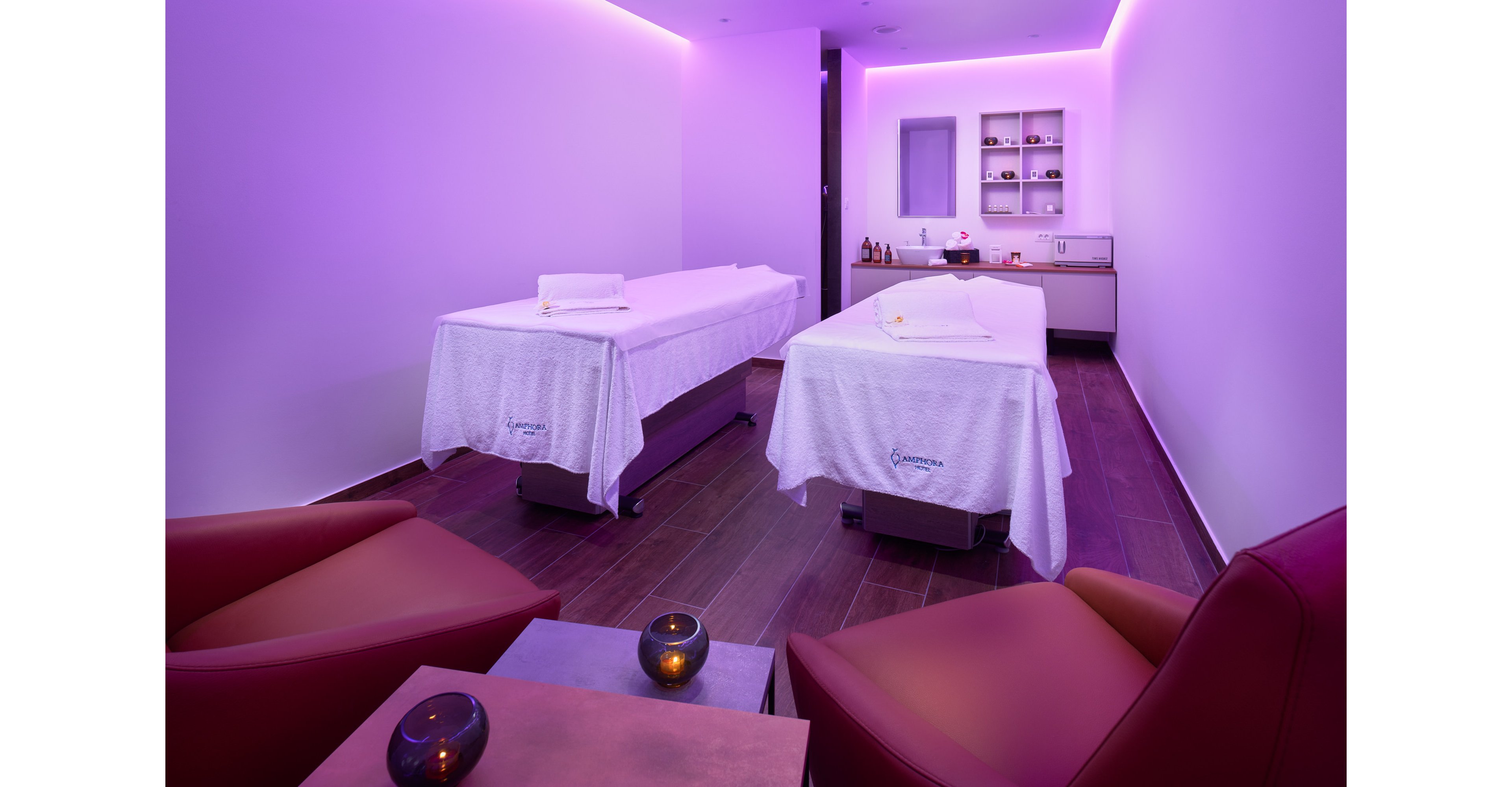 A contemporary spa treatment room features two massage tables neatly prepared with white linens. The space is illuminated by soft purple ambient lighting that creates a calming atmosphere. Shelves with towels, oils, and spa products are visible at the back of the room. Two comfortable lounge chairs and small tables with lit candles add to the relaxing, wellness-focused setting.