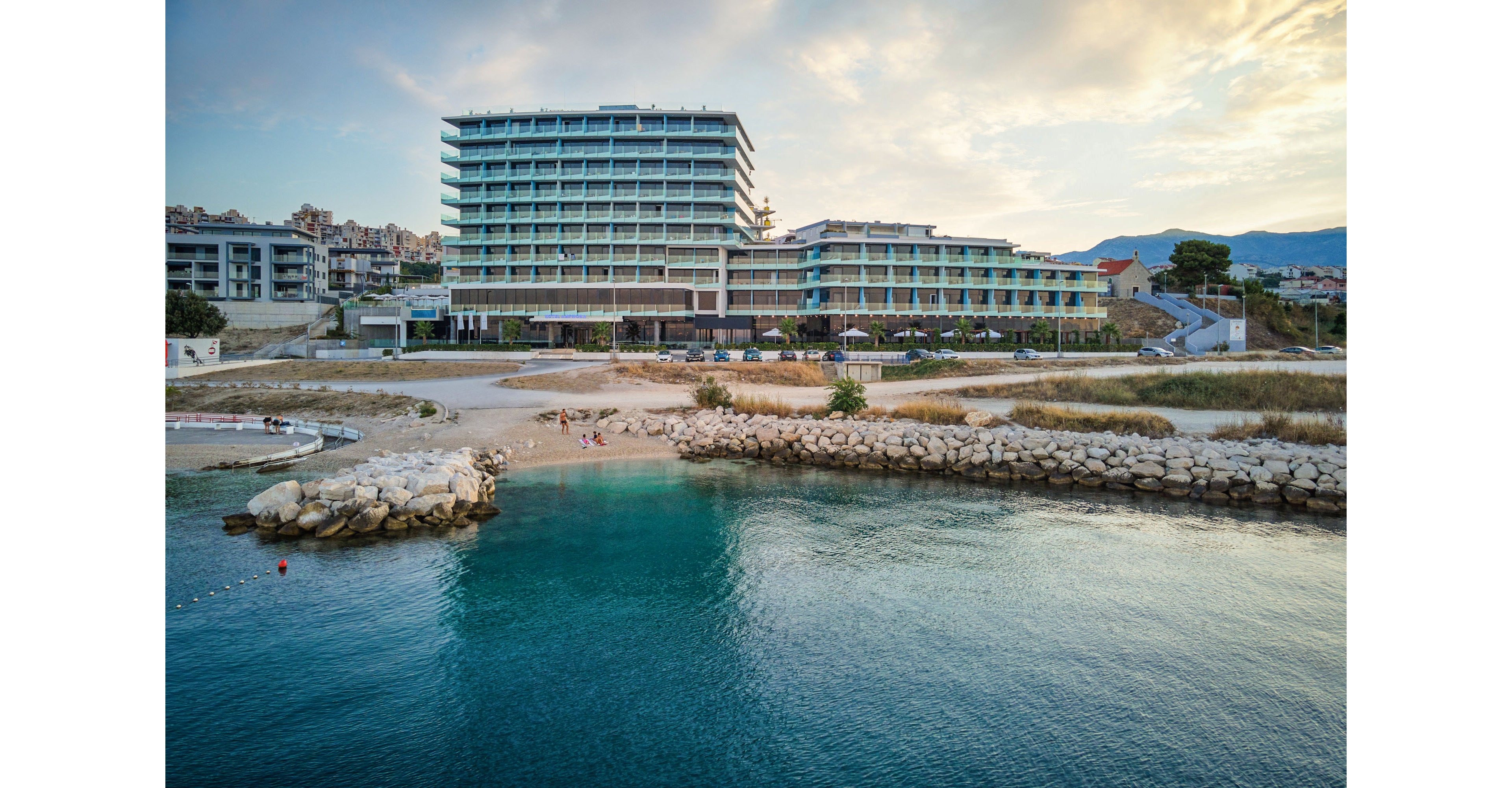 A large modern hotel complex stands along a quiet shoreline, facing a calm turquoise bay. The building features multiple stories with balconies and glass facades, surrounded by smaller coastal structures. A rocky breakwater and sandy beach separate the hotel from the clear water, where a few people can be seen near the shore. The scene is captured at sunset or early evening, with soft clouds and warm light in the sky. Distant hills and urban development frame the background, emphasizing a coastal resort setting.