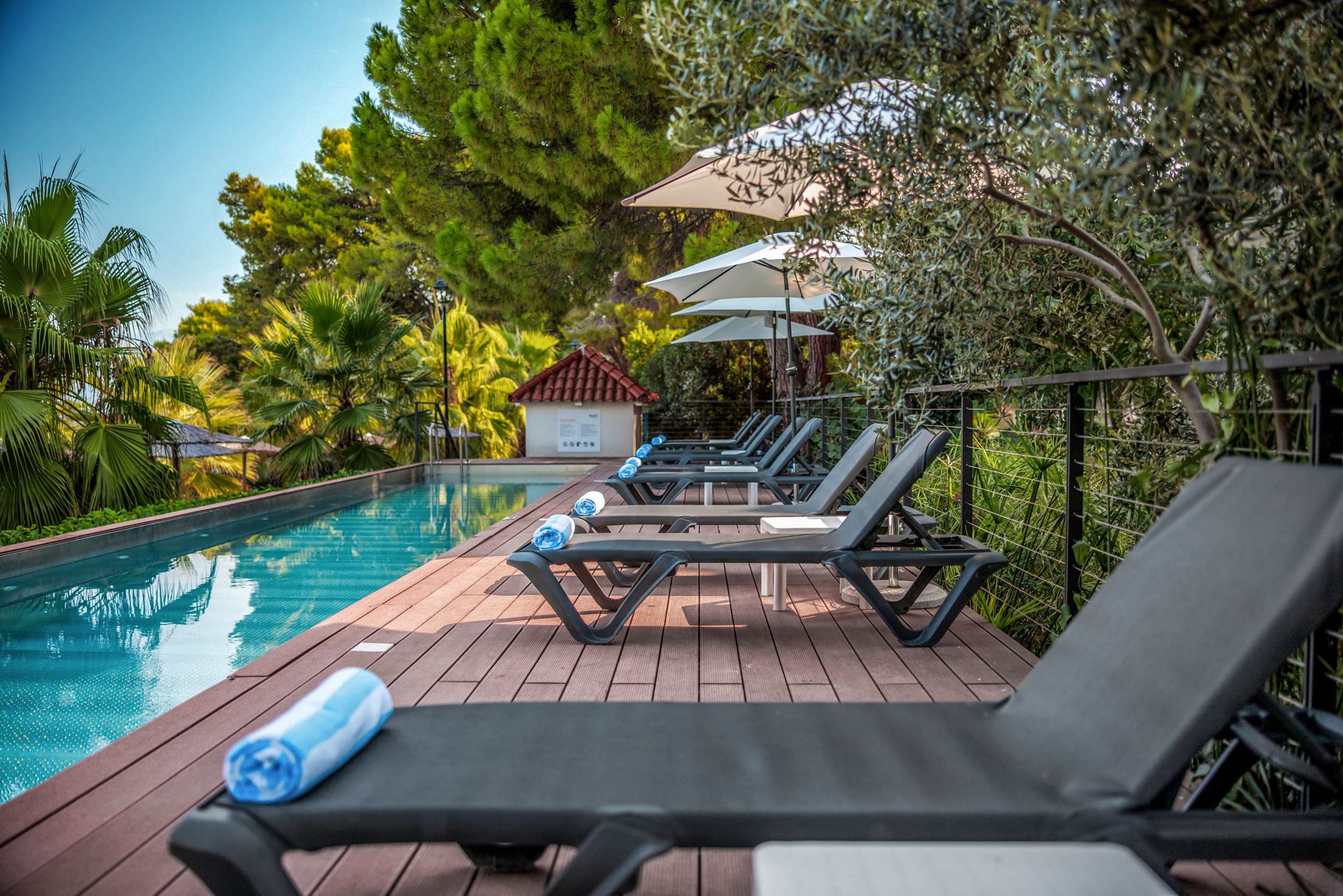 A modern outdoor swimming pool is lined with sleek sun loungers on a wooden deck. Rolled blue and white towels rest on the chairs, ready for guests. Lush green trees and plants surround the area, creating a private, resort-like atmosphere. Large umbrellas provide shade, enhancing the relaxing vacation setting.