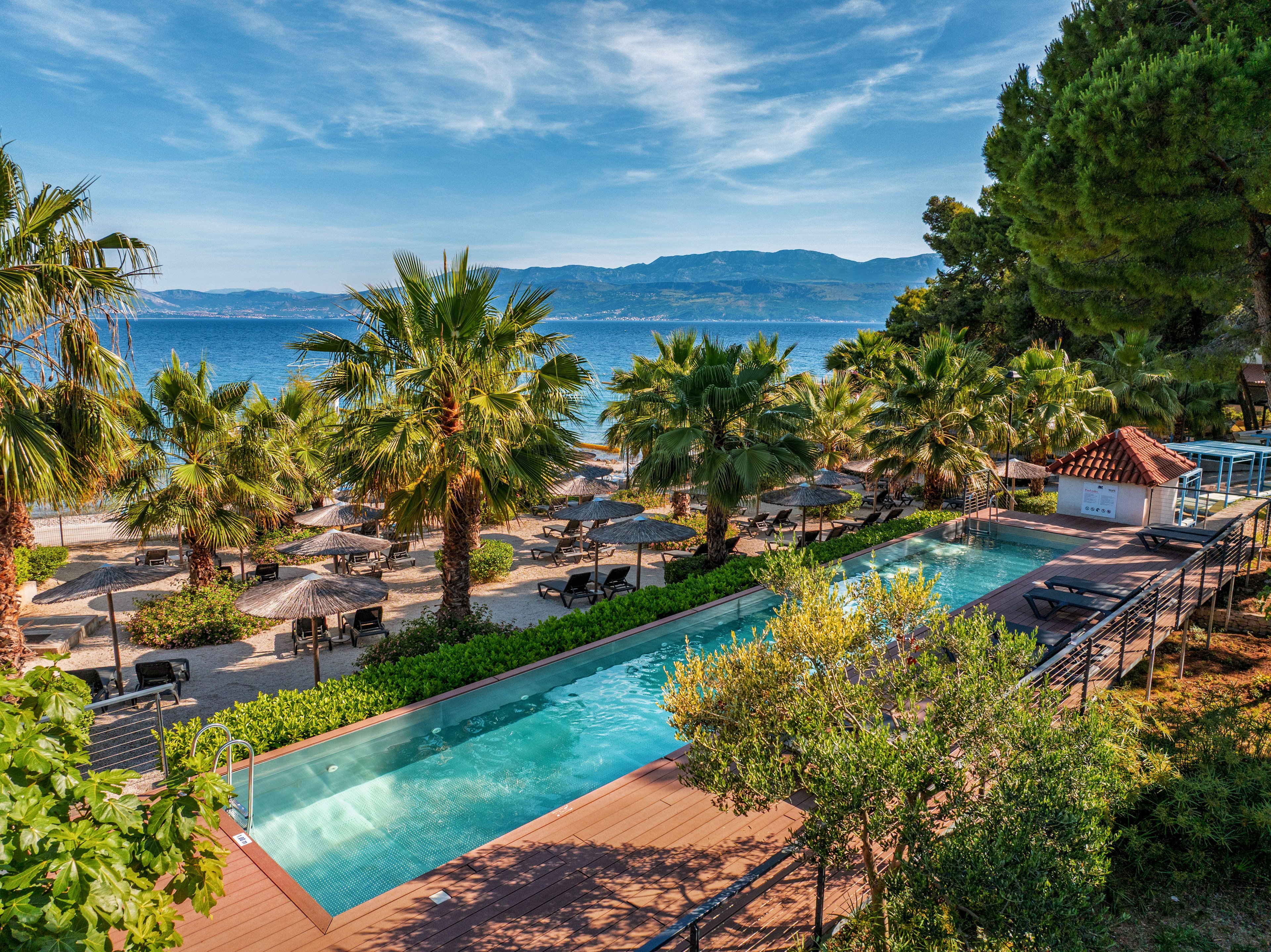 A long rectangular swimming pool overlooks a sandy beach lined with palm trees and sun loungers. Straw parasols and neatly arranged chairs create a relaxed vacation setting beside the calm blue sea. Greenery and landscaped paths frame the pool area, leading toward a small seaside structure. Distant mountains rise across the water under a bright, clear sky, enhancing the tranquil coastal atmosphere.