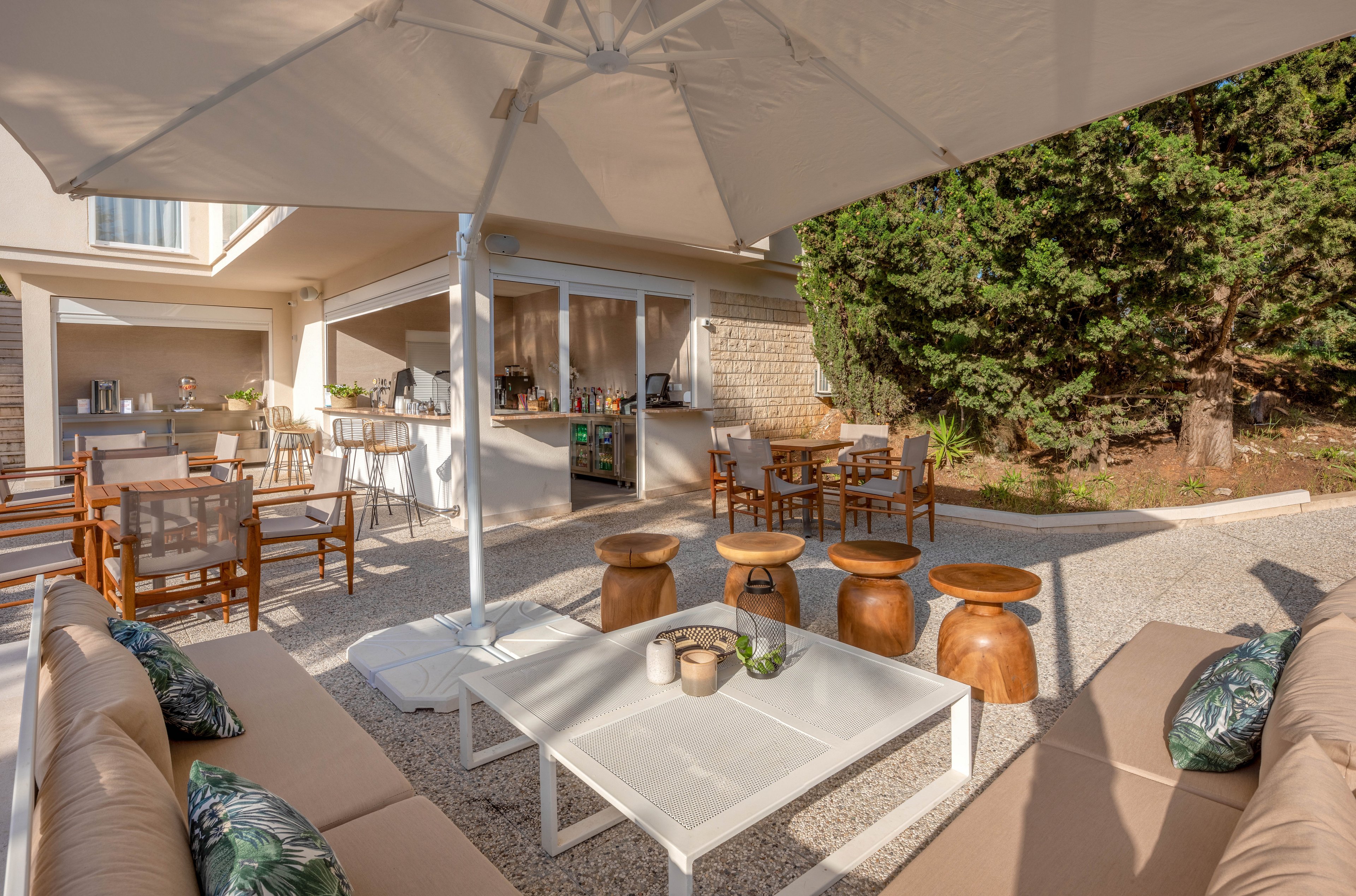 A spacious outdoor patio features contemporary seating arrangements under a large umbrella. Wooden chairs and stools surround tables, with a small bar or café area visible in the background. The setting is bright and inviting, with neutral tones and green accents from nearby trees. Cushions with leaf patterns add a touch of style to the beige sofas. No visible text or numbers are present in the image.