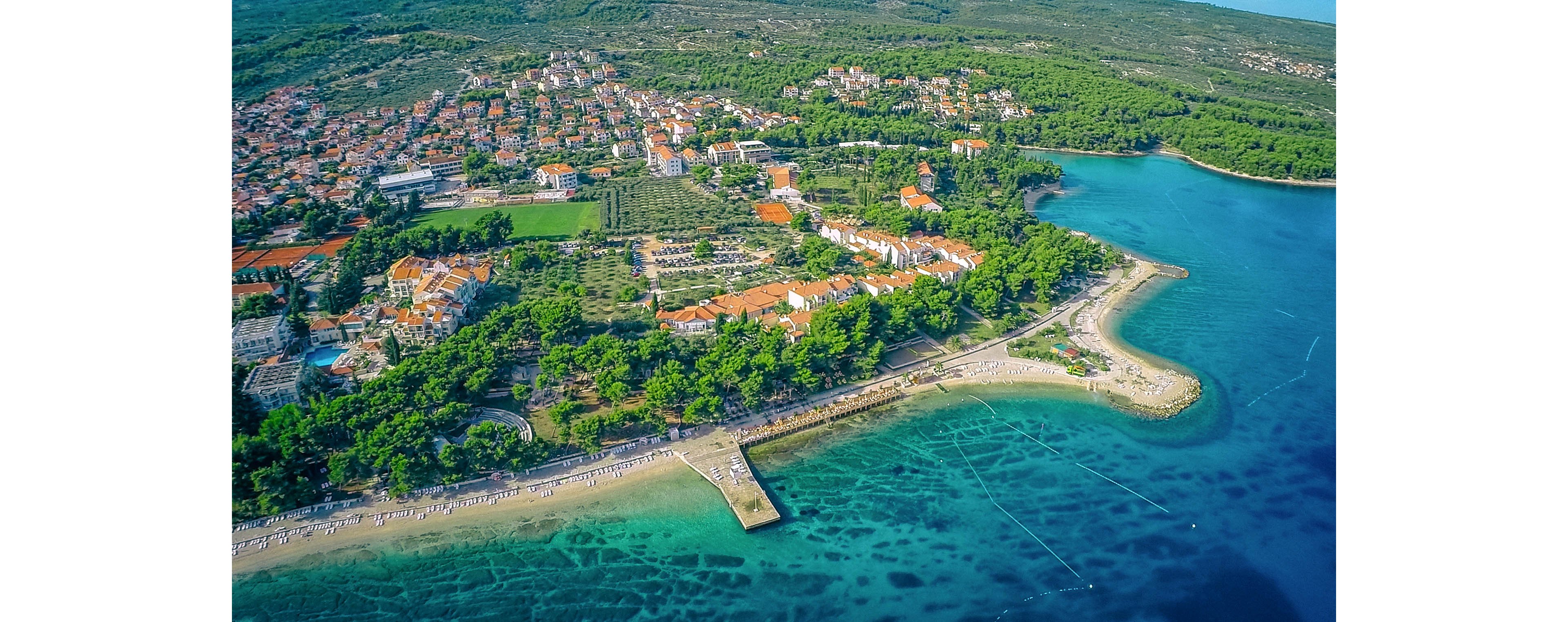This image captures a vibrant coastal town bordered by lush greenery and clear turquoise waters. The beachfront promenade is visible, lined with sunbathers and swimmers enjoying the sunny weather. Residential buildings with red-tiled roofs are nestled among trees, while the shoreline features a sandy beach and a pier extending into the sea. The overall mood is relaxed and inviting, with a bright, natural color palette.