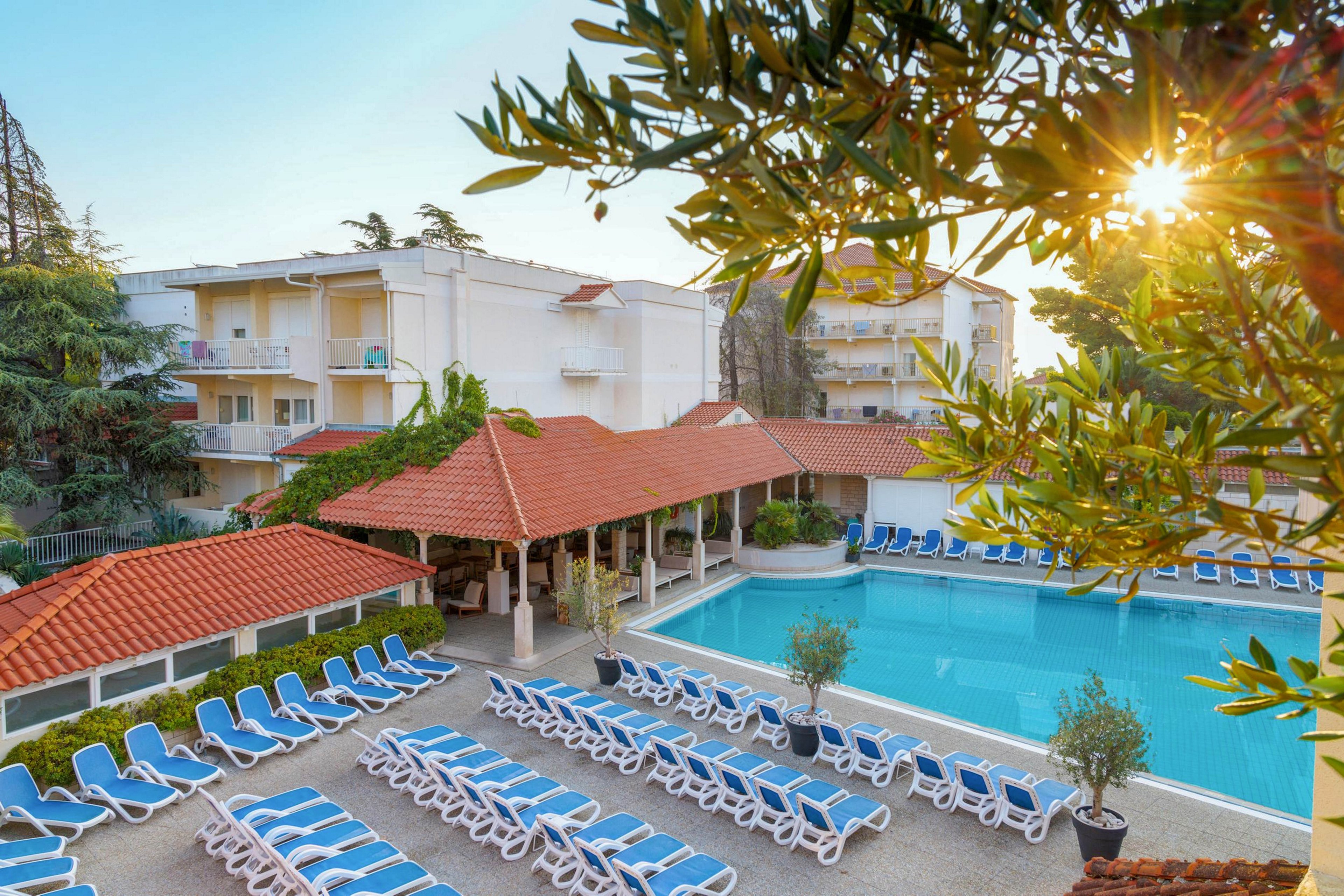 The image shows a spacious outdoor pool area at a resort, surrounded by rows of blue and white lounge chairs. The setting includes hotel buildings with balconies and a covered patio structure, all bathed in warm sunlight. The scene is tranquil, with lush greenery and a clear blue sky, evoking a relaxing vacation mood. No visible text or numbers are present in the image.
