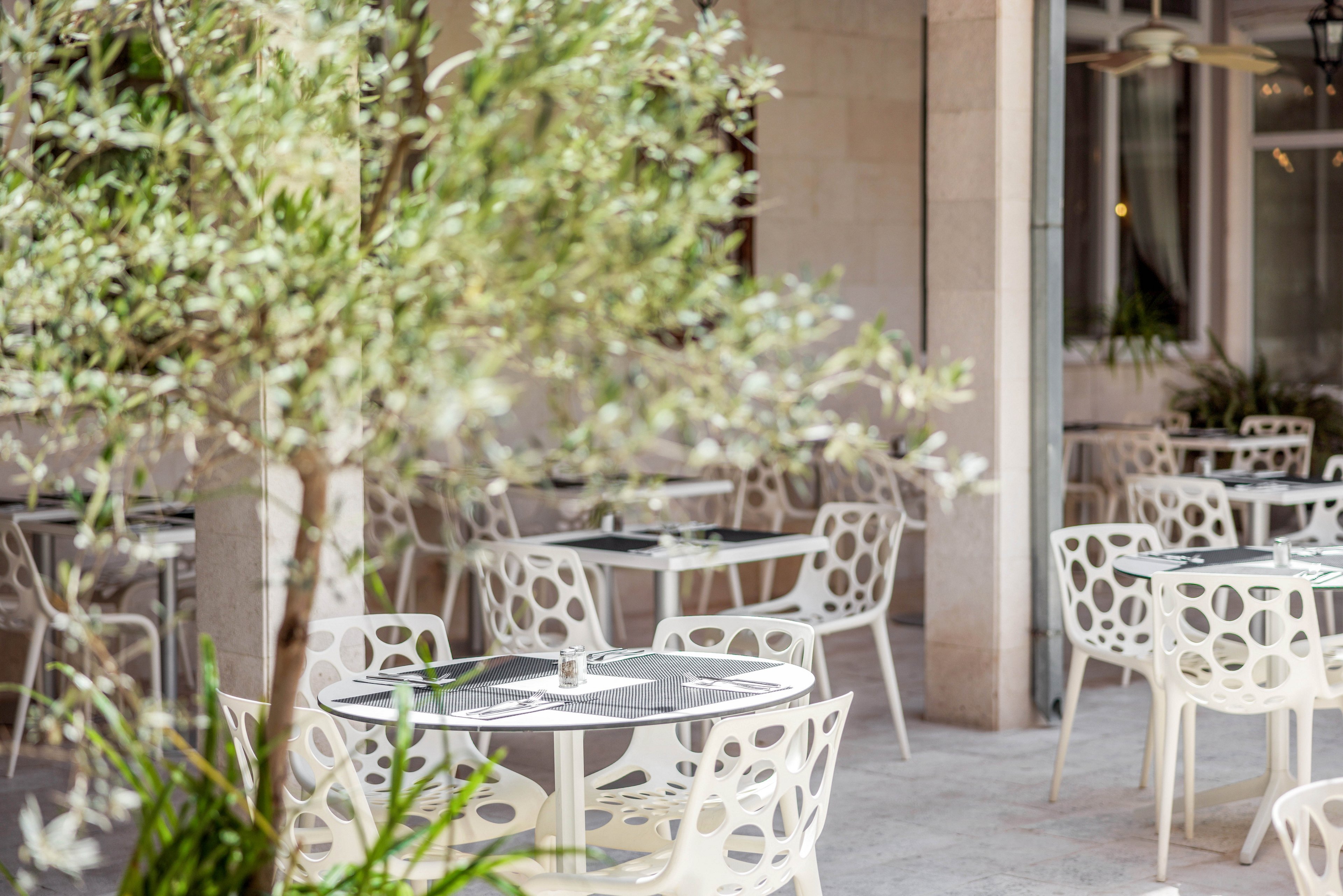 A stylish outdoor cafe features contemporary white chairs and round tables set on a stone patio. The setting is bright and airy, with leafy plants adding a touch of greenery to the scene. The tables are neatly arranged, each set with cutlery and glasses, suggesting a welcoming atmosphere for dining. The overall mood is relaxed and inviting, ideal for casual gatherings.