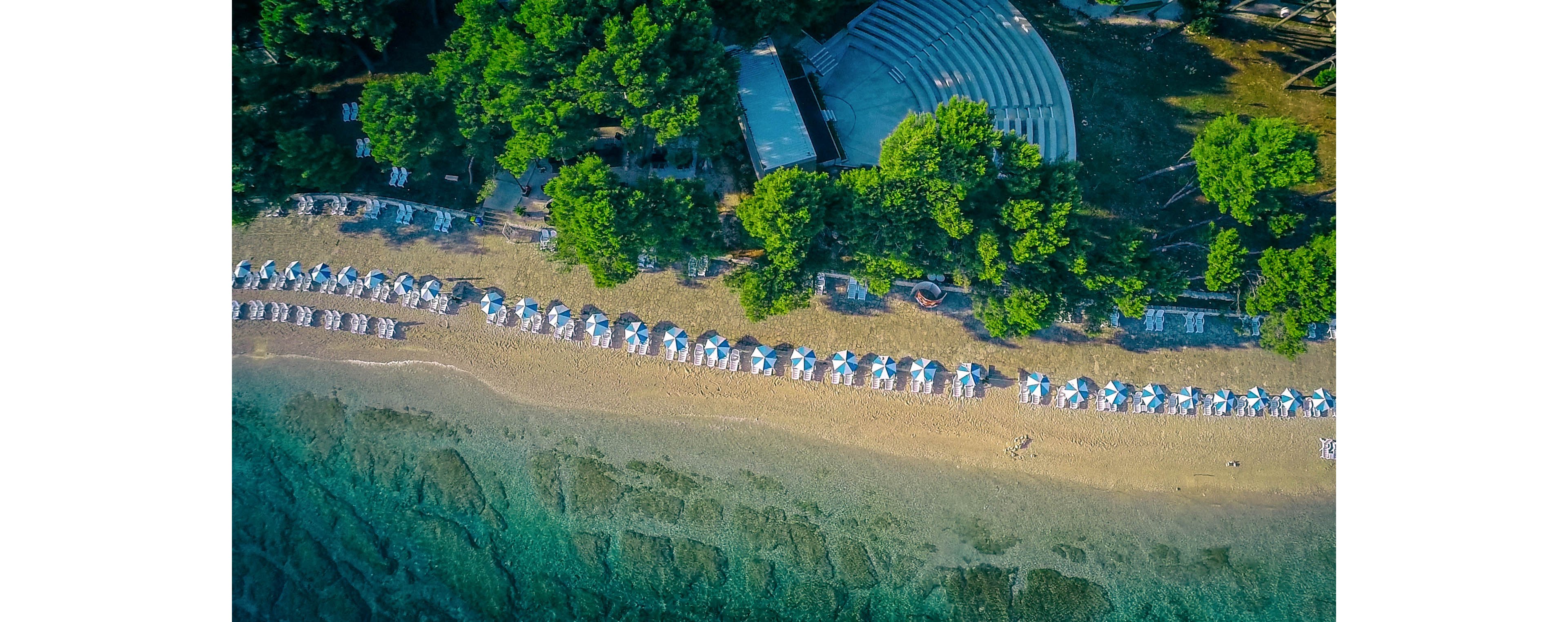 This aerial photograph captures a scenic beach lined with rows of white and blue umbrellas and sun loungers. The shoreline meets clear turquoise water, while dense green trees provide shade and a natural border. A large building structure is partially visible among the trees, suggesting a resort or recreational facility. The overall mood is tranquil and inviting, with a vibrant summer palette.